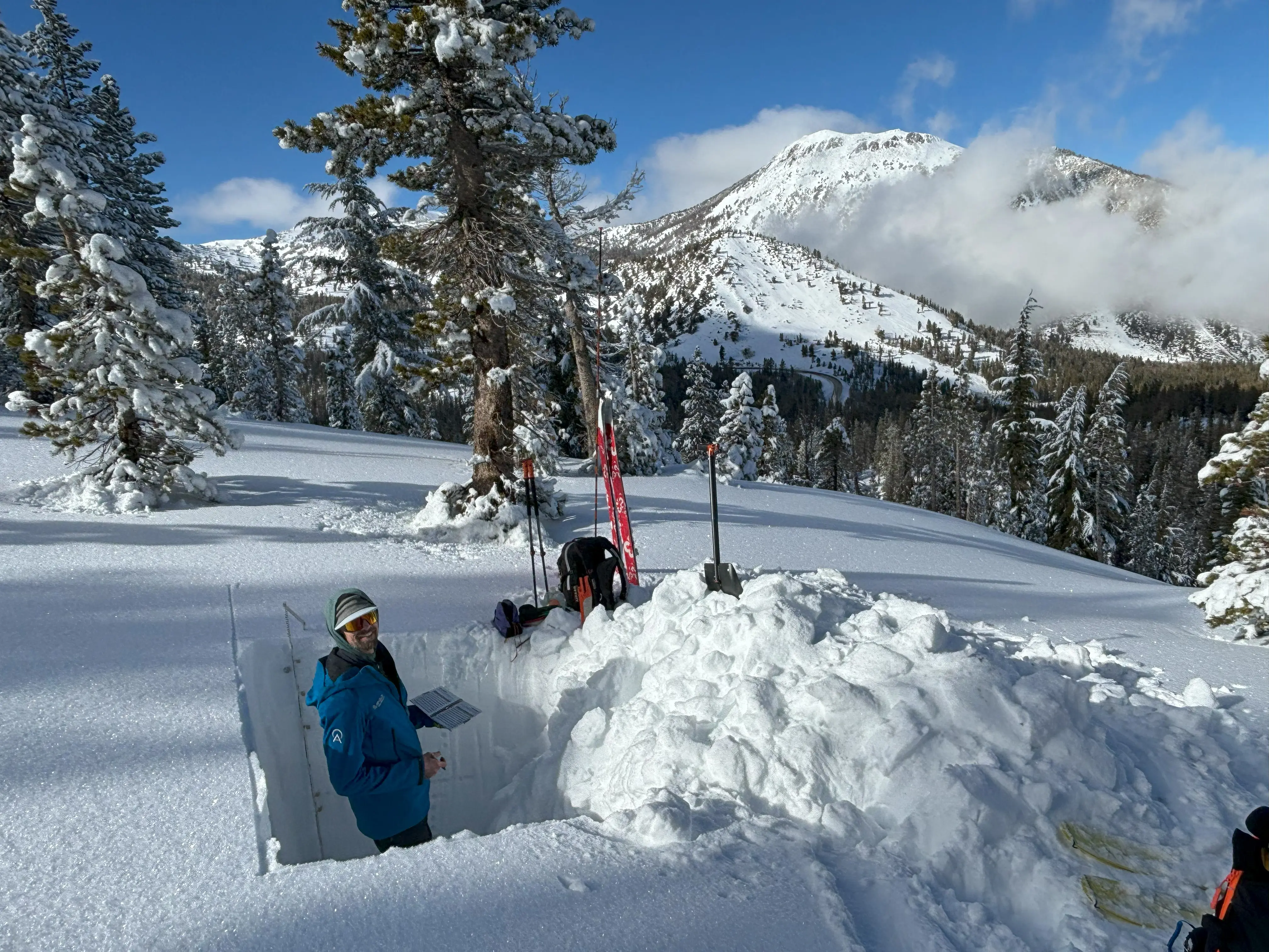 Two people are examining snow in a dug-out pit on a snowy mountain slope. Skis and poles are nearby. One person is recording data on a laptop, while the other is inside the pit. Pine trees and a snow-covered mountain are in the background. Mt. Rose Ski Tahoe