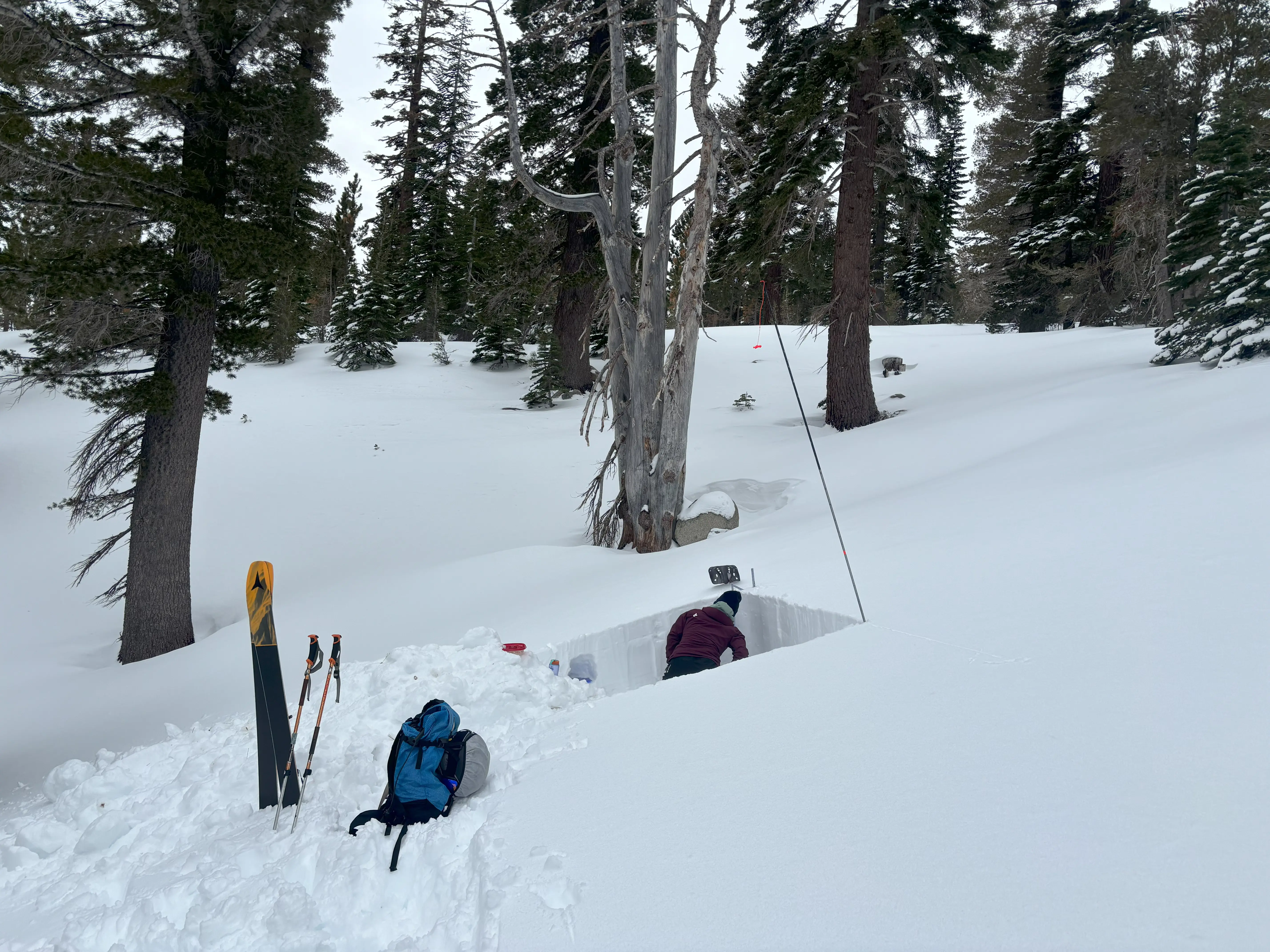 A person is kneeling down inside a dug-out rectangular snow pit, surrounded by trees. In the snow nearby are a pair of skis, ski poles, and a backpack. Snow blankets the landscape, suggesting a winter setting. Mt. Rose Ski Tahoe
