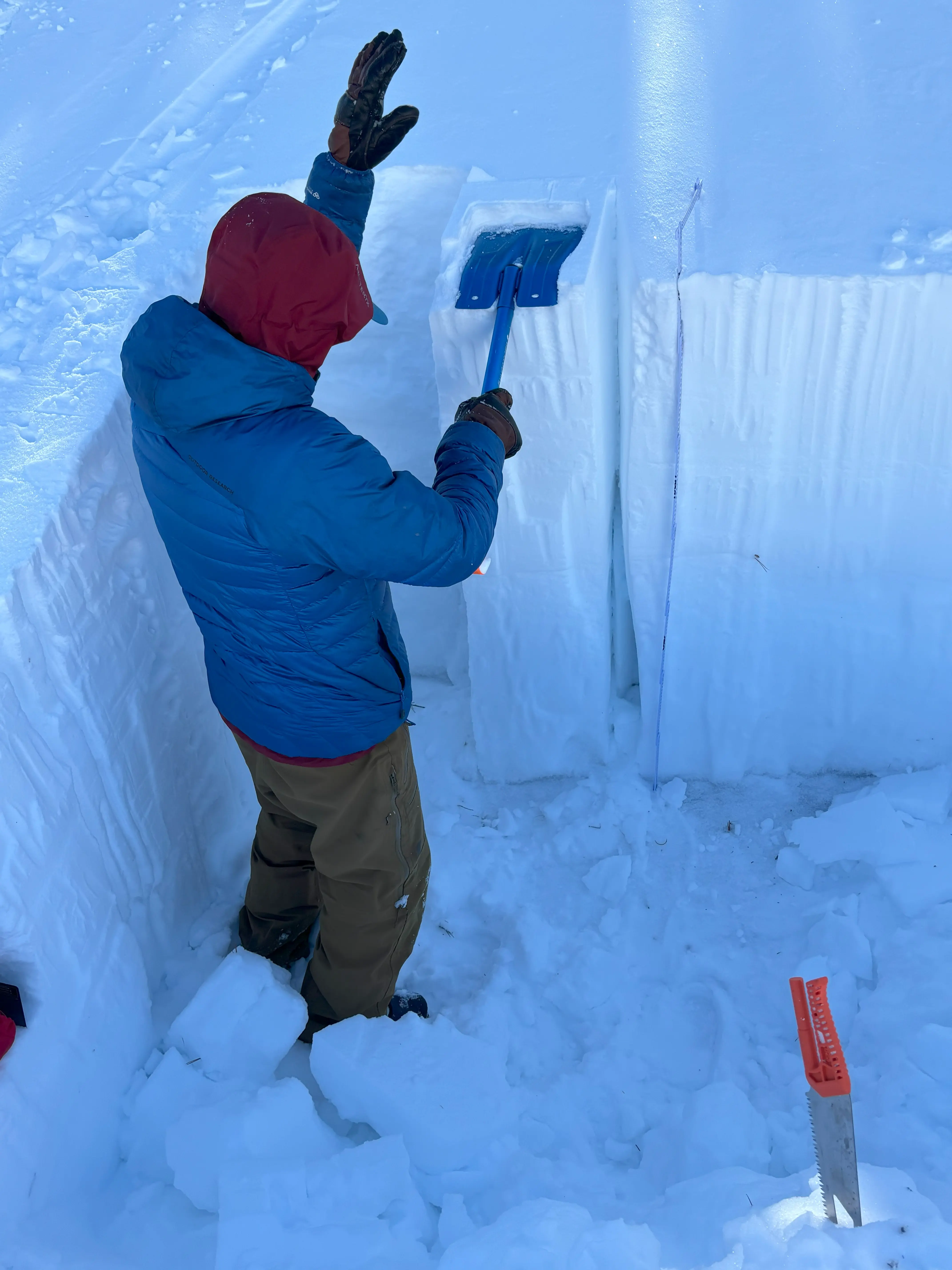 A person in a red hooded jacket and brown pants digs a trench in the snow. They hold a blue shovel, standing in an area surrounded by cut snow blocks. The scene suggests preparation for an avalanche study or snow profile. Mt. Rose Ski Tahoe