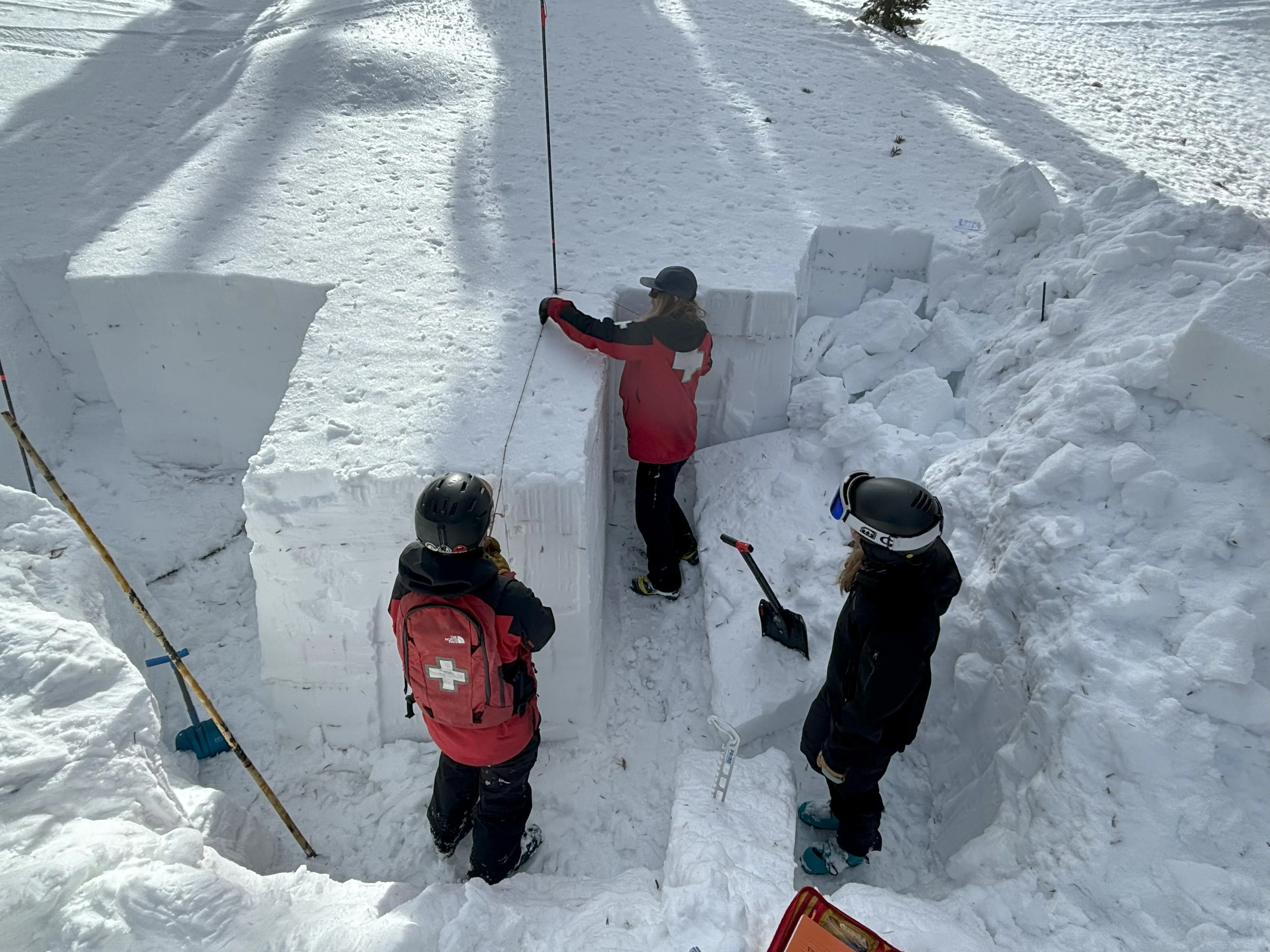 Three people in winter gear and helmets, two wearing red with white cross symbols, are studying a cut section of snow. Shovels and ski poles are nearby, and the scene suggests an avalanche training or snow study in a snowy landscape. Mt. Rose Ski Tahoe