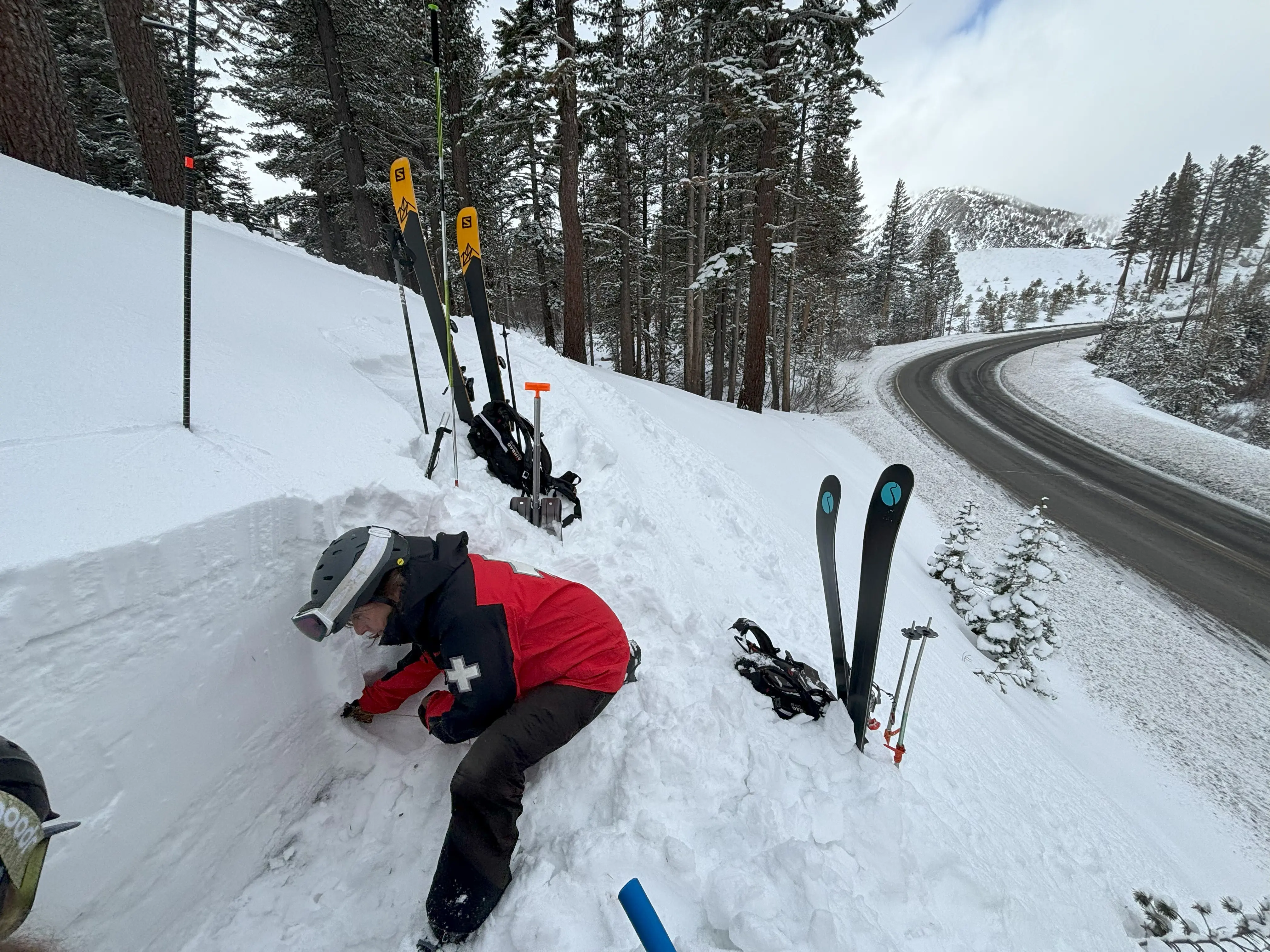 A person in a red and black jacket is digging into a snowpack on a snowy slope near a curved road. Skis and poles are stuck upright in the snow beside them. Trees and snow-covered mountains are in the background. Mt. Rose Ski Tahoe