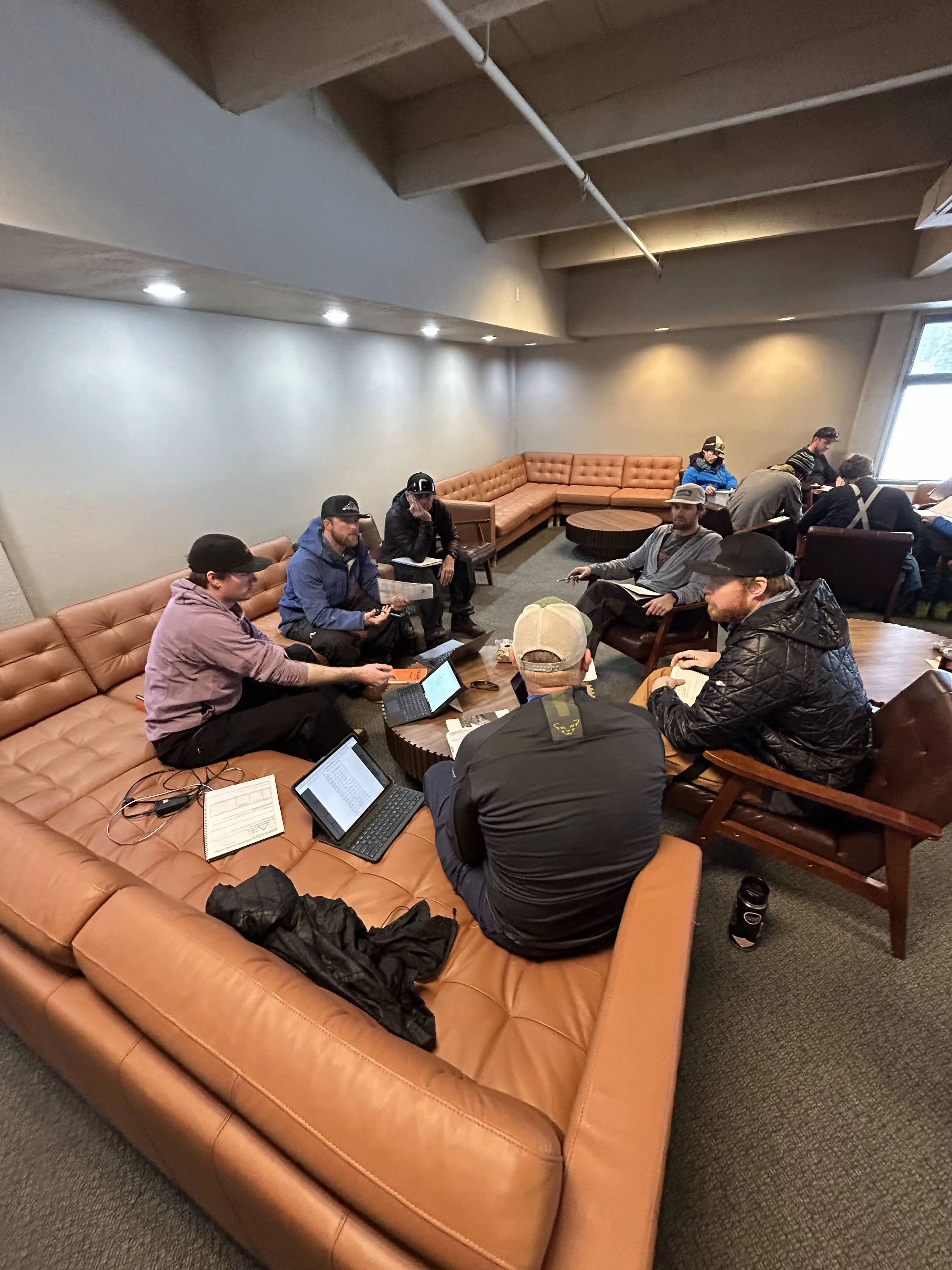 A group of people sit in a circle on brown leather couches in a lounge area, having a discussion. Laptops and notebooks are open on the coffee tables in front of them. The room has a cozy atmosphere with dim lighting. Mt. Rose Ski Tahoe