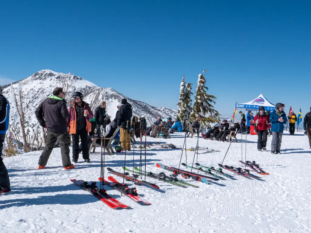 A snowy mountain scene with several people gathered, some standing and others sitting. Skis and poles are lined up on the snow. A tent in the background reads "Demo Center." Clear blue sky and a snow-covered peak complete the view. Mt. Rose Ski Tahoe A snowy mountain scene with several people gathered, some standing and others sitting. Skis and poles are lined up on the snow. A tent in the background reads "Demo Center." Clear blue sky and a snow-covered peak complete the view. Mt. Rose Ski Tahoe
