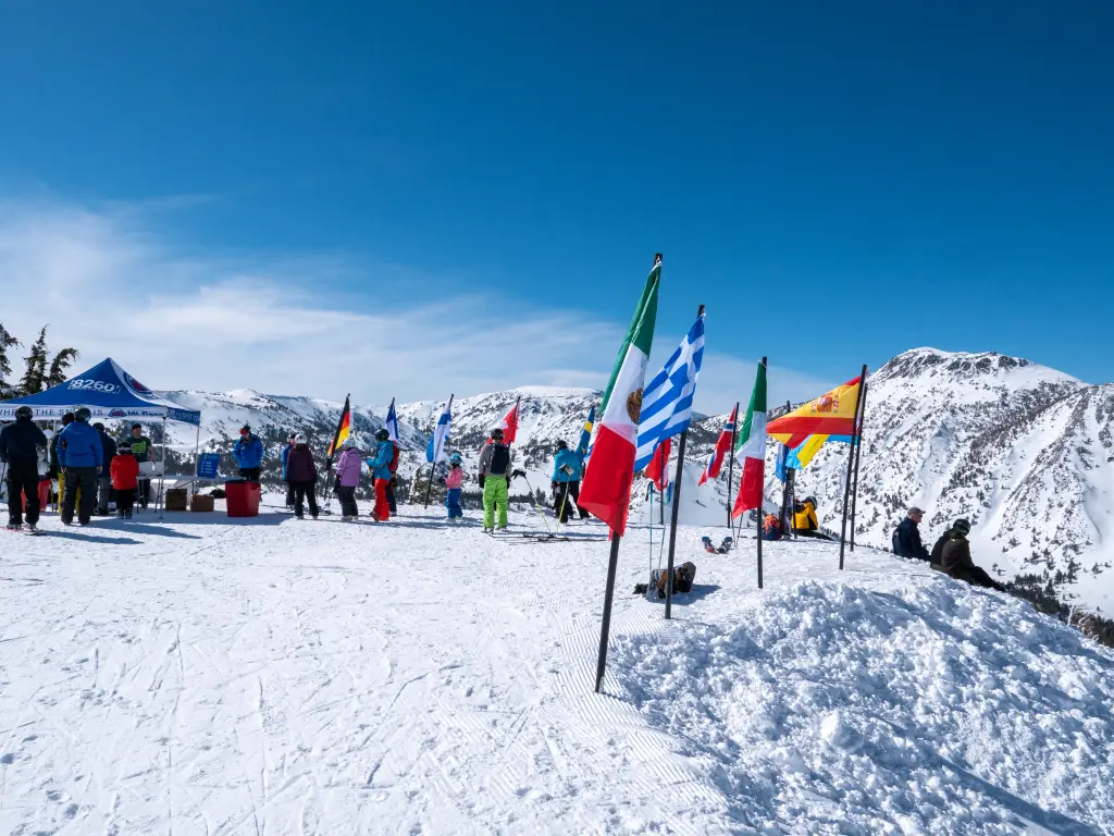 A snowy mountain peak with various national flags, including Italy, Greece, and Colombia, waving in the wind. People in winter gear are standing and interacting near a blue tent. Snow-covered mountains are visible under a clear blue sky. Mt. Rose Ski Tahoe A snowy mountain peak with various national flags, including Italy, Greece, and Colombia, waving in the wind. People in winter gear are standing and interacting near a blue tent. Snow-covered mountains are visible under a clear blue sky. Mt. Rose Ski Tahoe