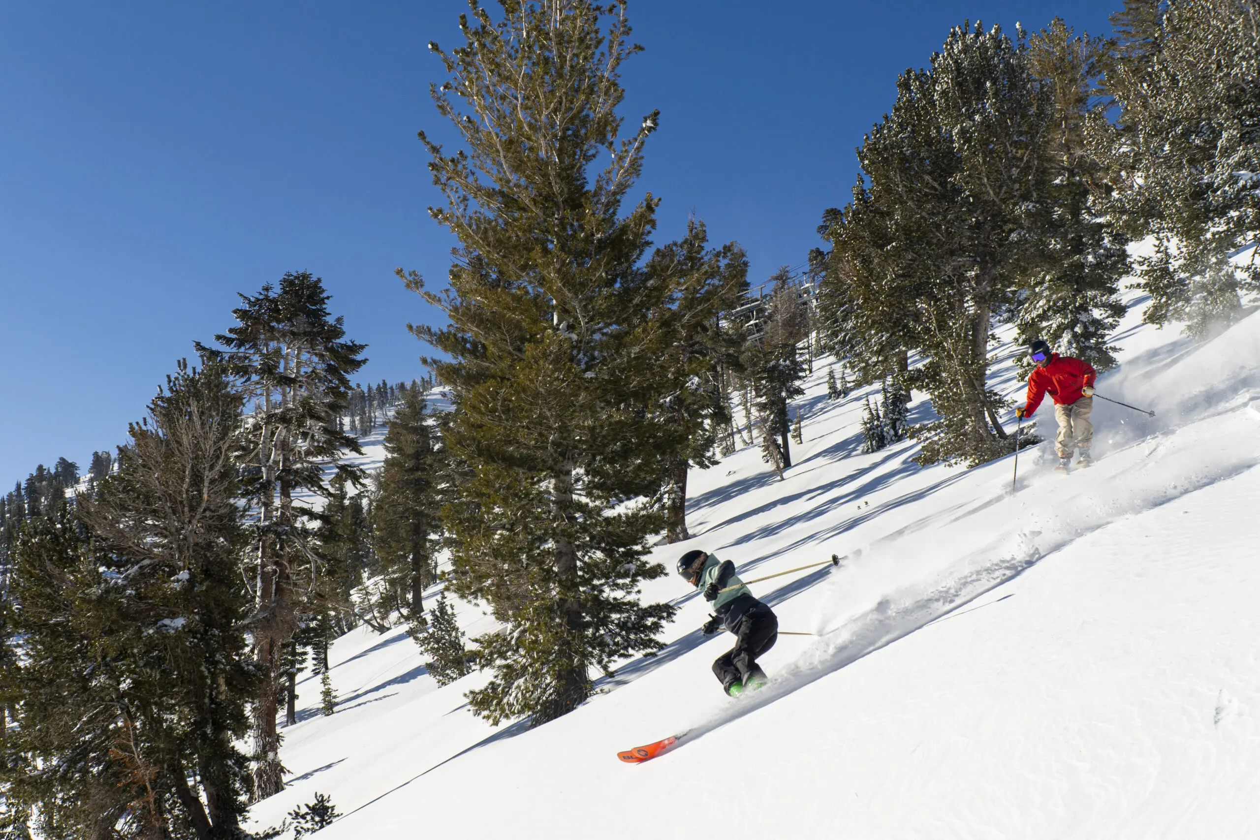 A skier in a light green jacket and black helmet moves swiftly downhill at Ski Tahoe, kicking up a spray of snow. They are surrounded by evergreen trees under a clear blue sky. Mt. Rose Ski Tahoe