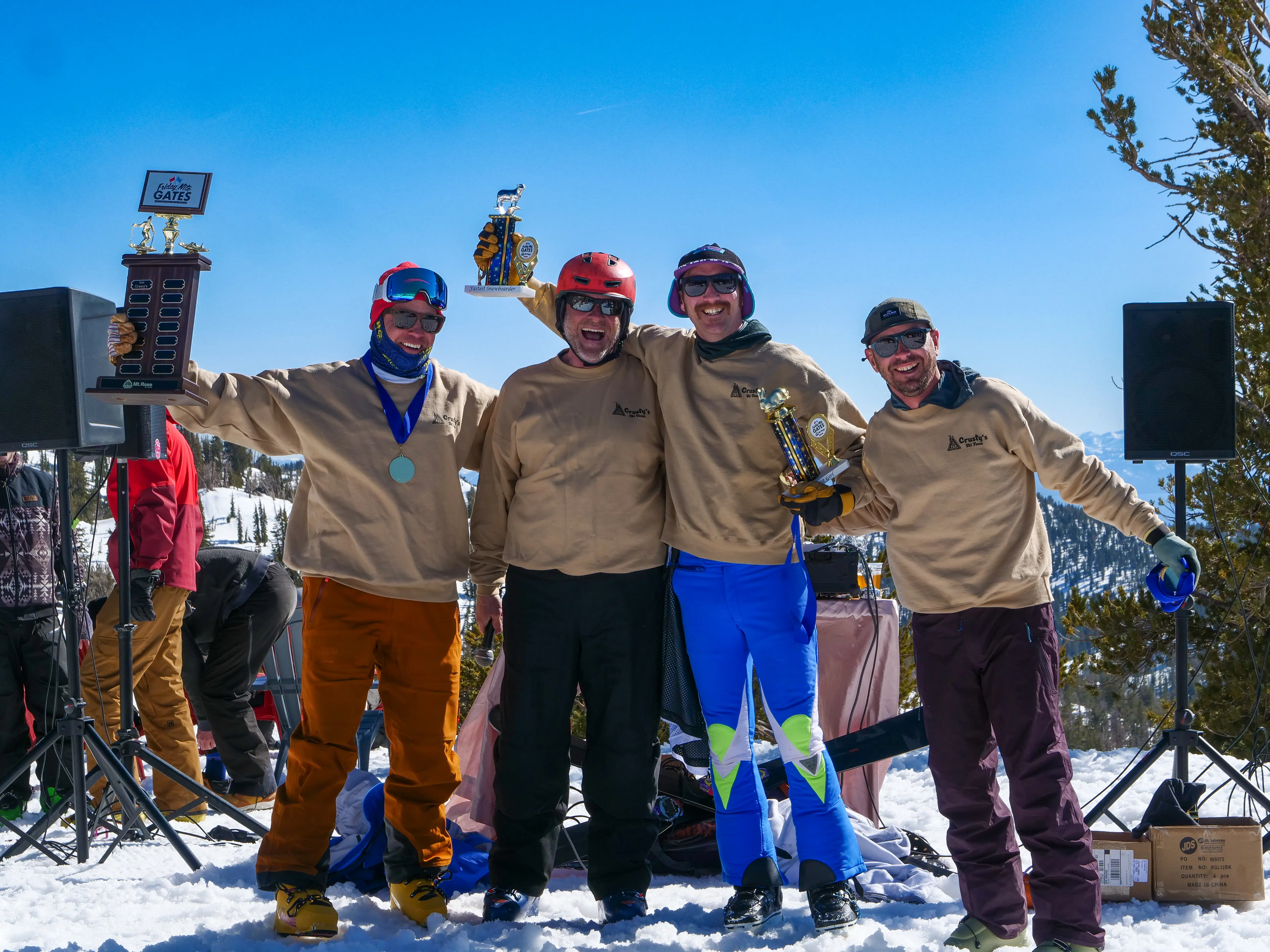 Four people in matching beige shirts and colorful snow pants stand on snow, smiling and holding Ski Racing trophies. A blue sky and mountains are in the background, with ski gear and speakers nearby. Mt. Rose Ski Tahoe