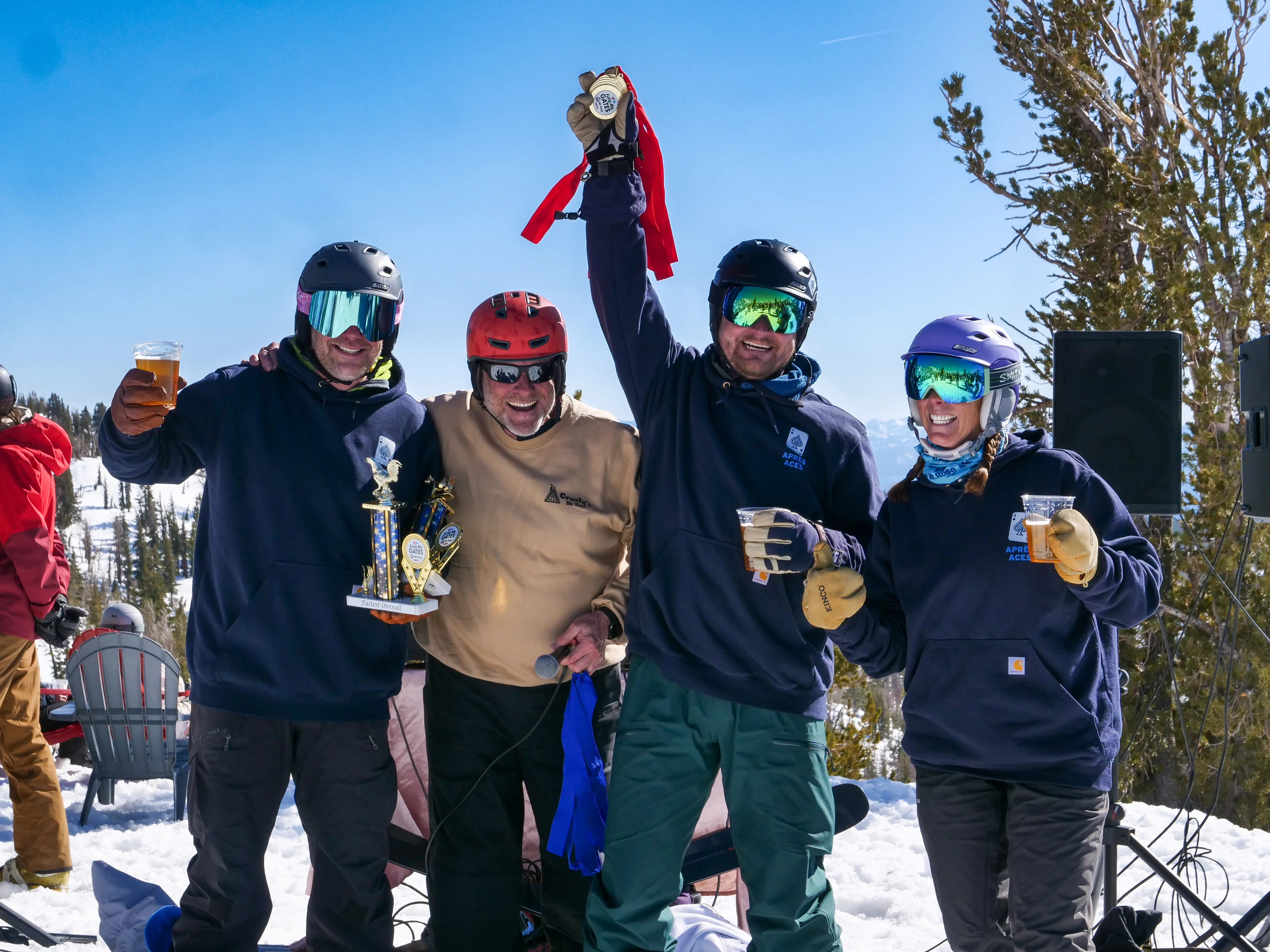Four people in winter gear stand in snow, smiling and celebrating. One man holds up a medal, another holds a trophy, and others raise drinks. Trees and snowy mountains are in the background under a clear blue sky. Mt. Rose Ski Tahoe
