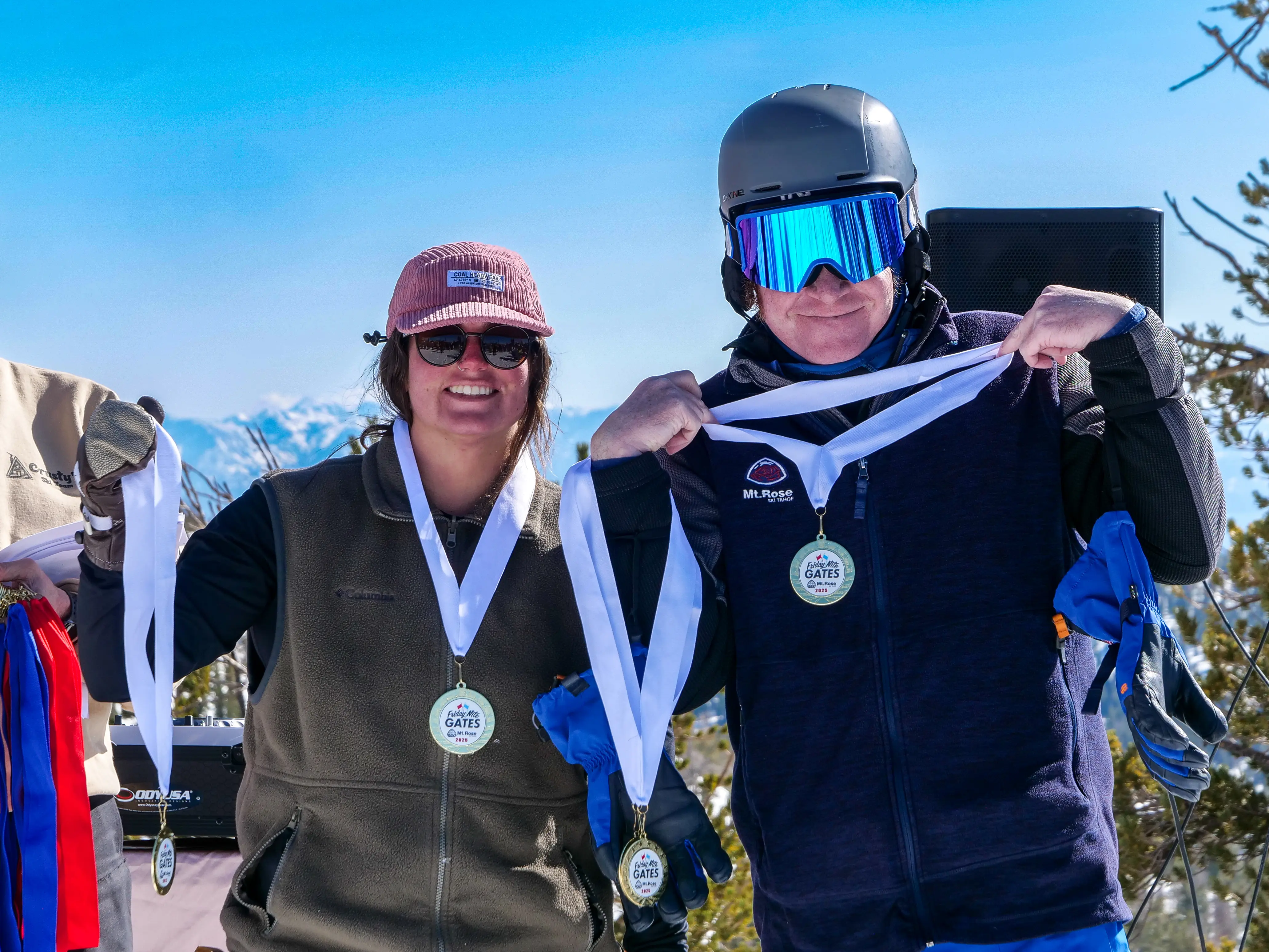 Two people in winter gear and sunglasses smile outdoors, each holding up a medal with a snowy mountain landscape behind them. They appear to be celebrating an achievement on a sunny day. Mt. Rose Ski Tahoe