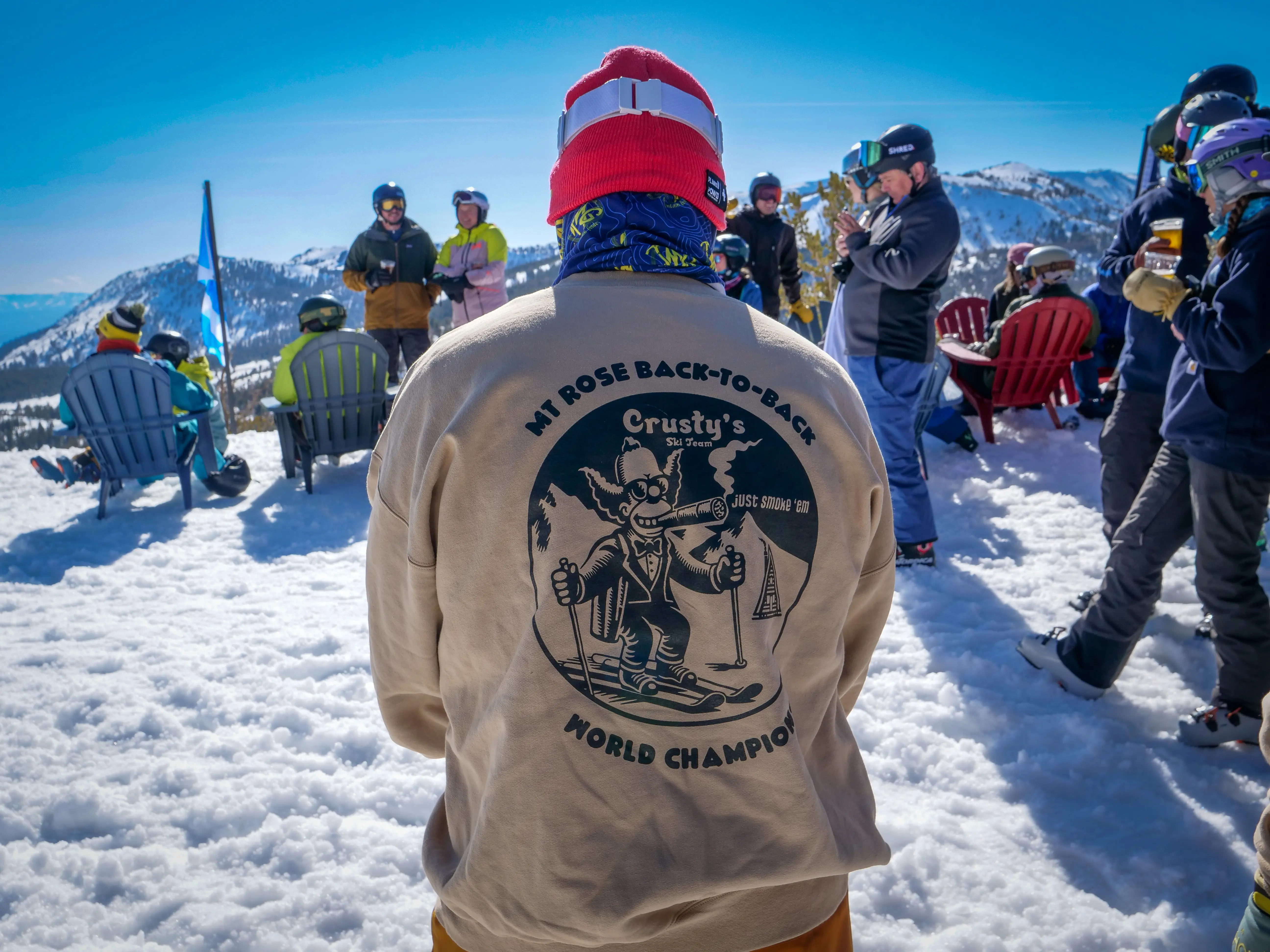 A person wearing a beige jacket with a cartoon skier and “Mt. Rose Back-to-Back World Champion” stands in snowy mountains, facing a group of people sitting and standing outdoors in winter gear under a clear blue sky. Mt. Rose Ski Tahoe