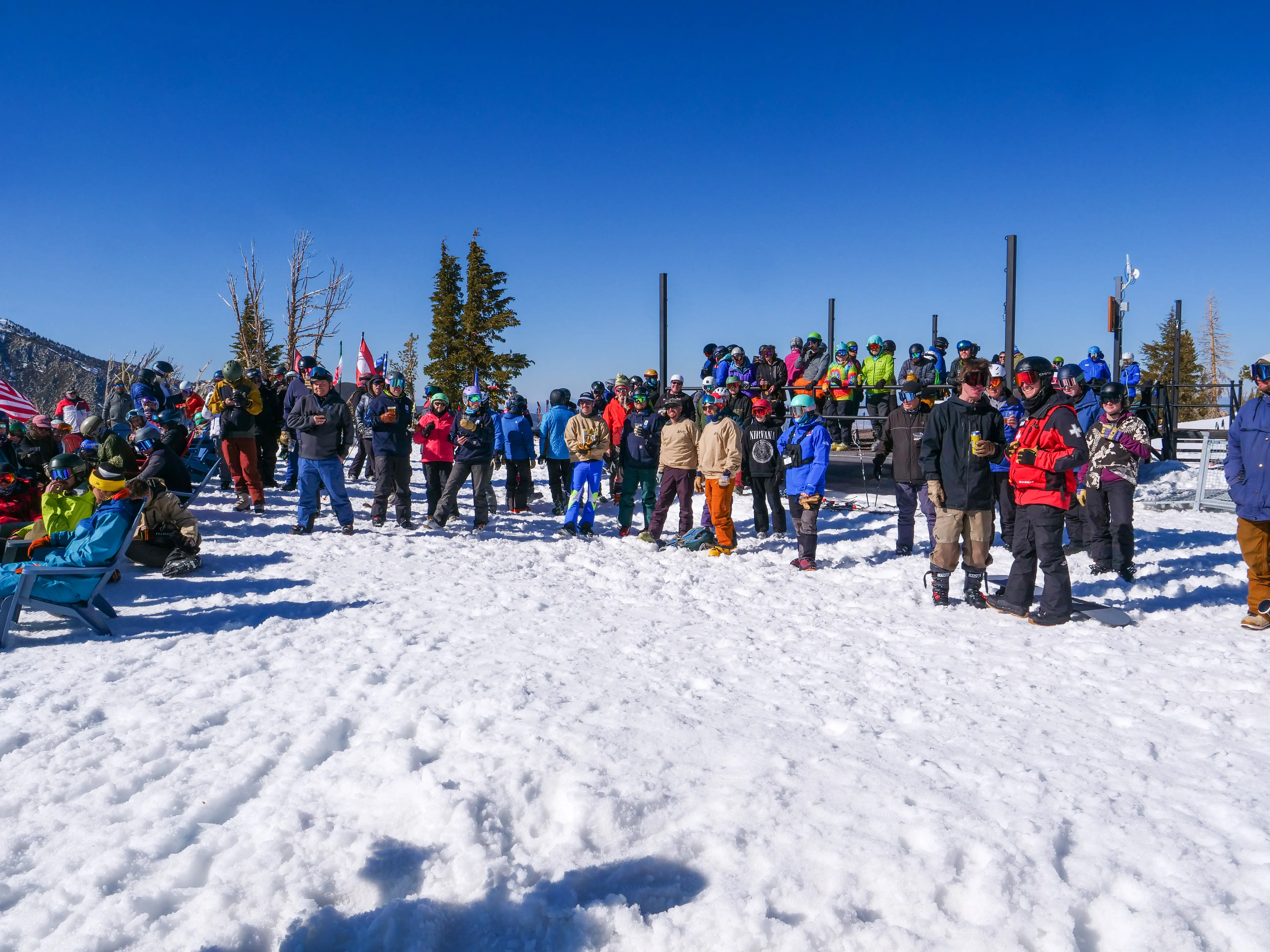 A large group of people in colorful winter clothing and helmets gather on a snowy mountain under a clear blue sky, likely waiting to ski or snowboard. Trees and ski poles are visible in the background. Mt. Rose Ski Tahoe