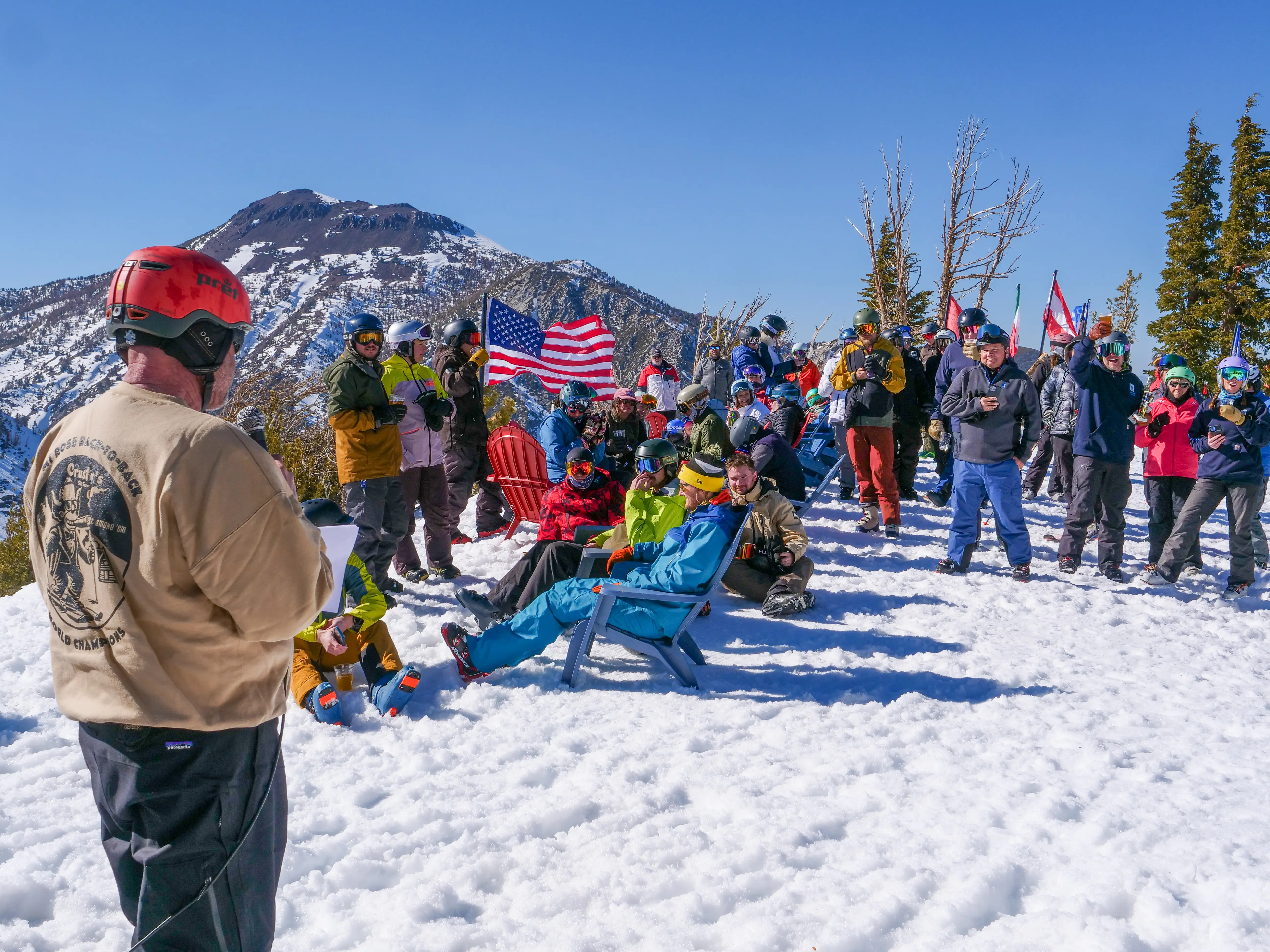 A group of people in winter clothing gathers on a snowy mountain slope, some holding flags, as a man in a helmet and tan shirt speaks to them. Snow-covered peaks and trees are visible in the background under a clear blue sky. Mt. Rose Ski Tahoe