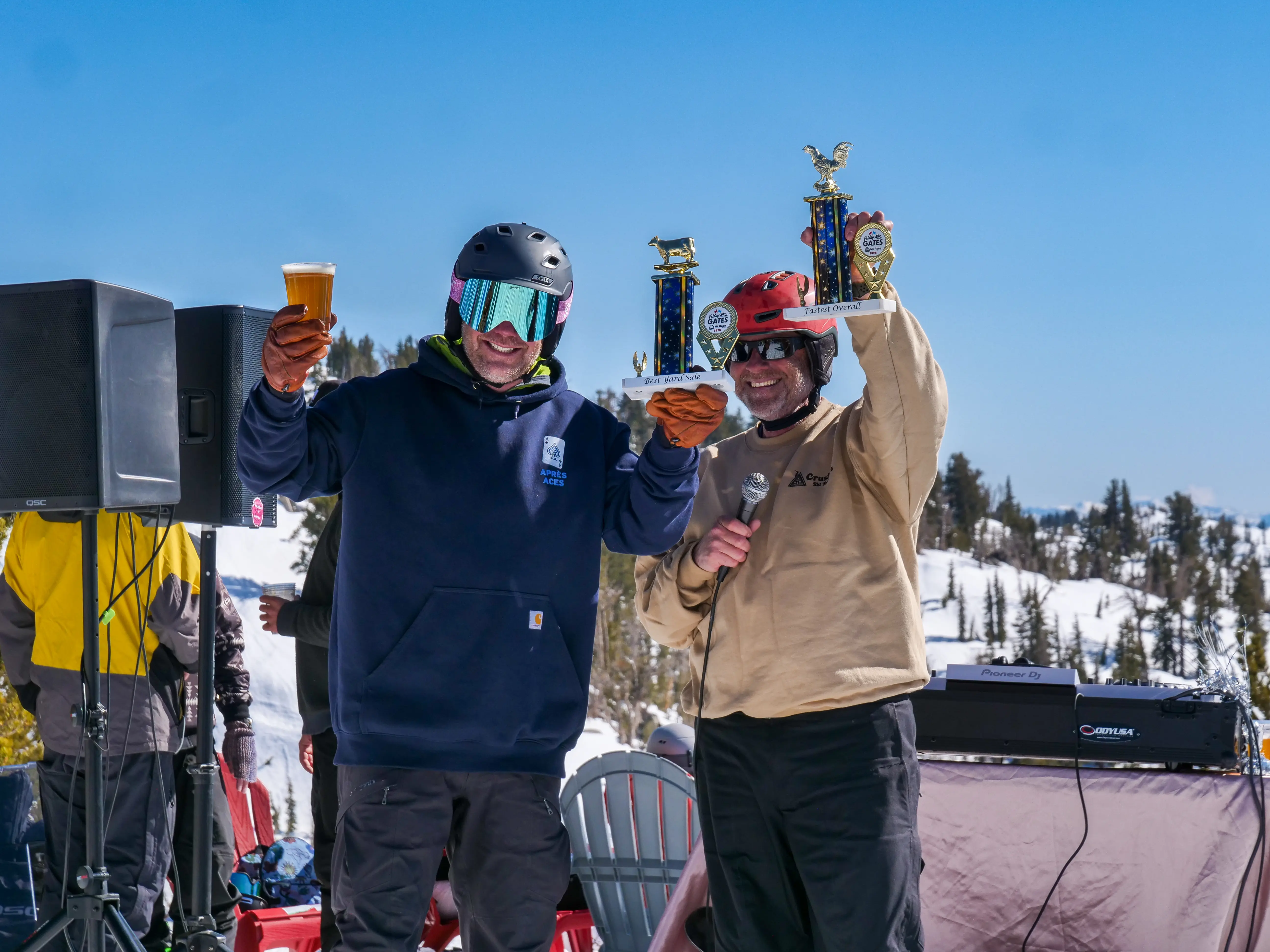 Two people in ski gear celebrate outdoors; one holds a beer and the other holds trophies and a microphone. Snowy mountains and a clear blue sky are in the background. Mt. Rose Ski Tahoe