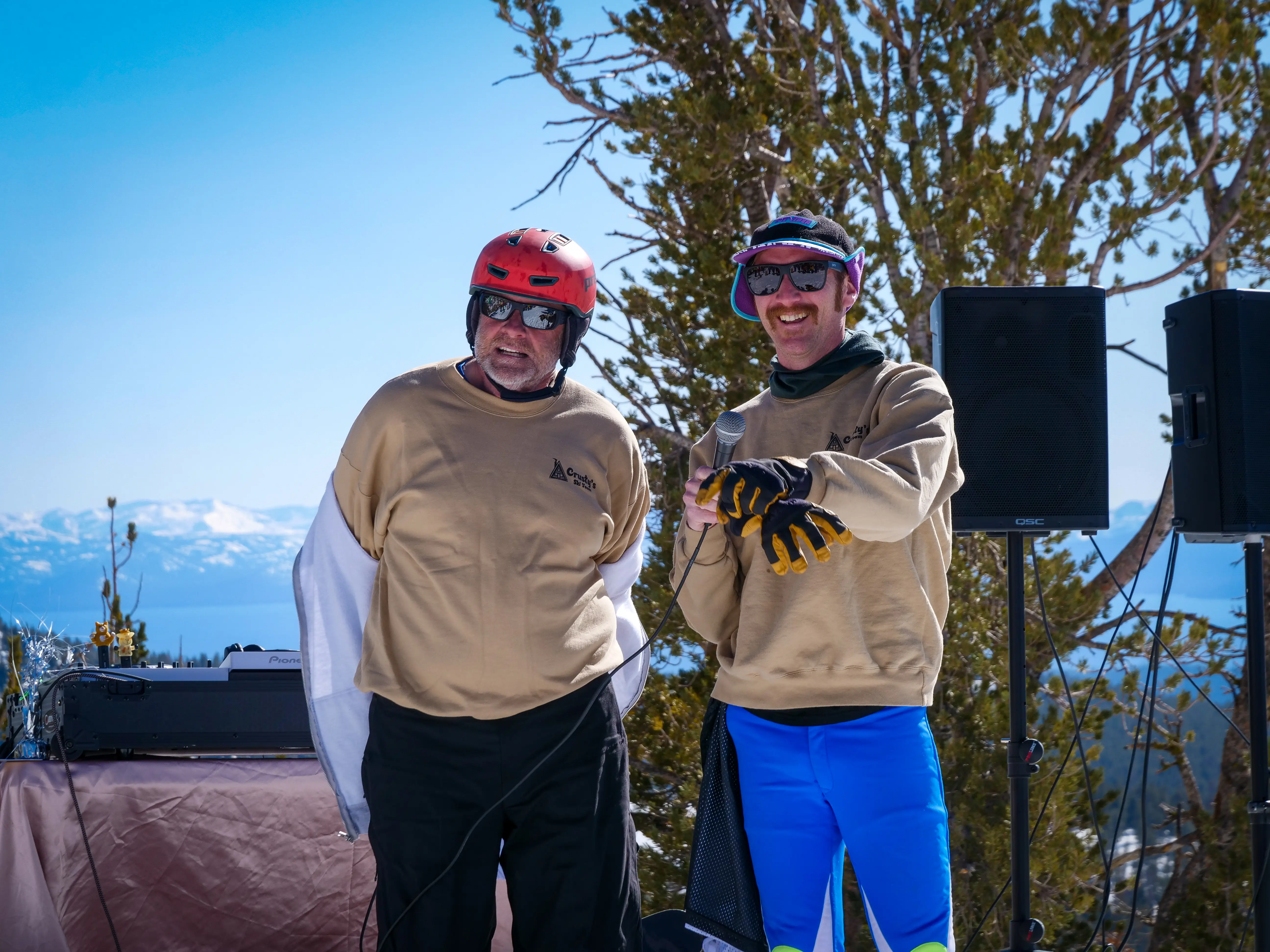 Two men in tan sweatshirts stand outdoors in front of trees and speakers; one wears a red helmet and sunglasses, the other wears a cap, pink sunglasses, blue pants, and yellow gloves. Snow-capped mountains are visible in the background. Mt. Rose Ski Tahoe