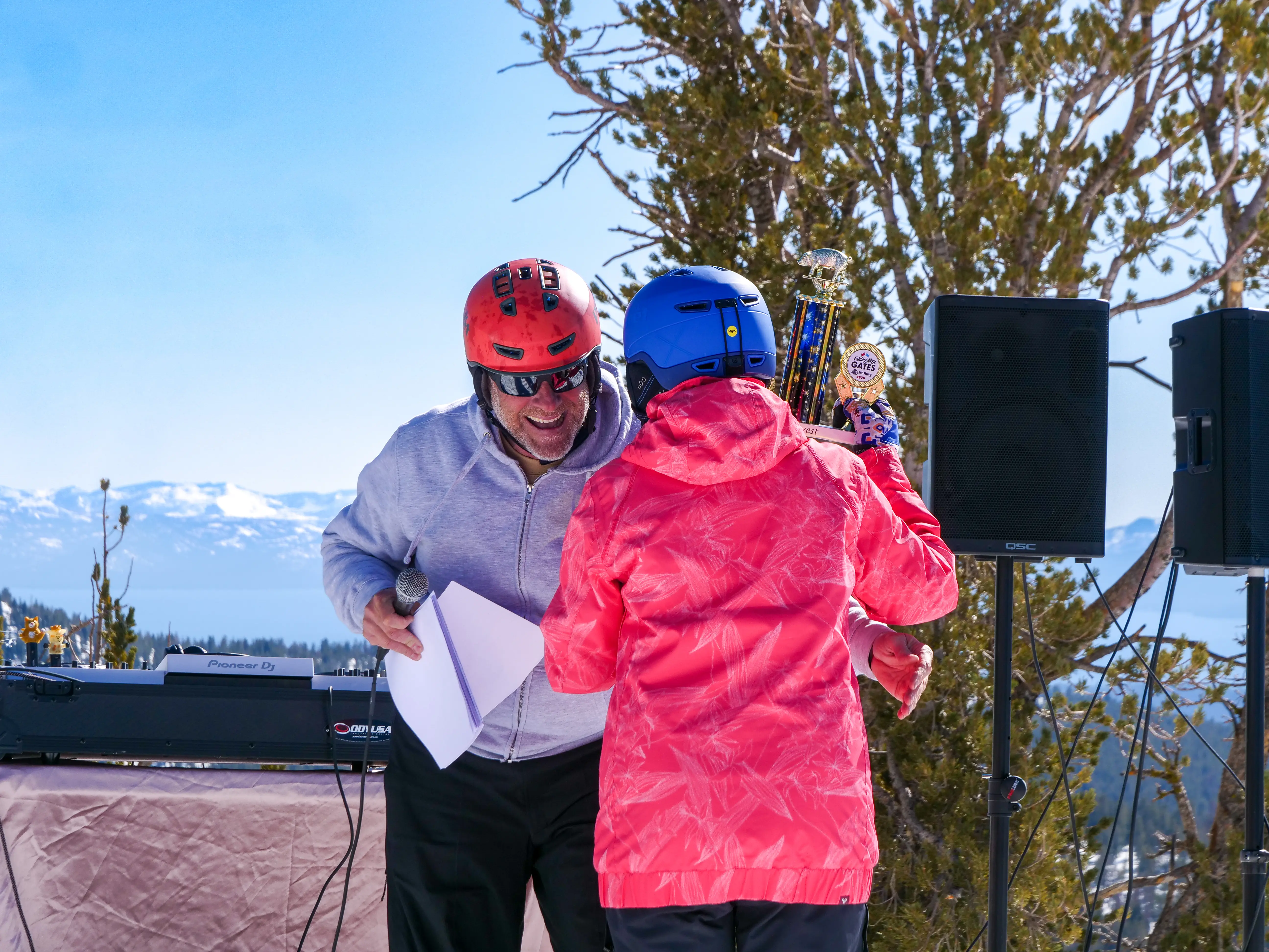 Two people in helmets and winter jackets stand outdoors, one holding a trophy and the other smiling with a sheet of paper. Snow-capped mountains and trees are visible in the background. Mt. Rose Ski Tahoe