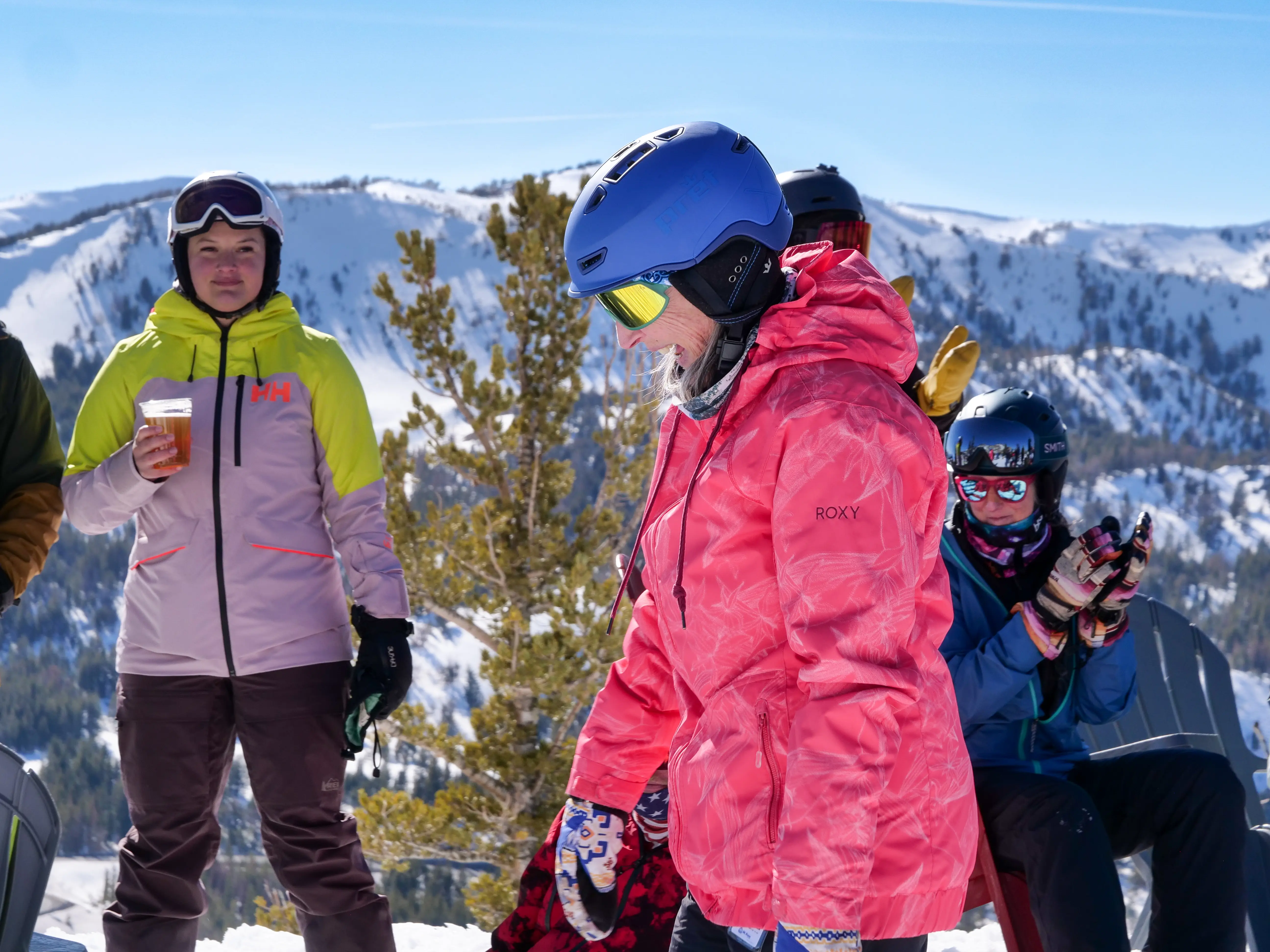 People in colorful ski gear stand and sit outdoors in the snow, with snowy mountains in the background. One person holds a drink, and another wears a pink jacket and blue helmet, looking down. Mt. Rose Ski Tahoe