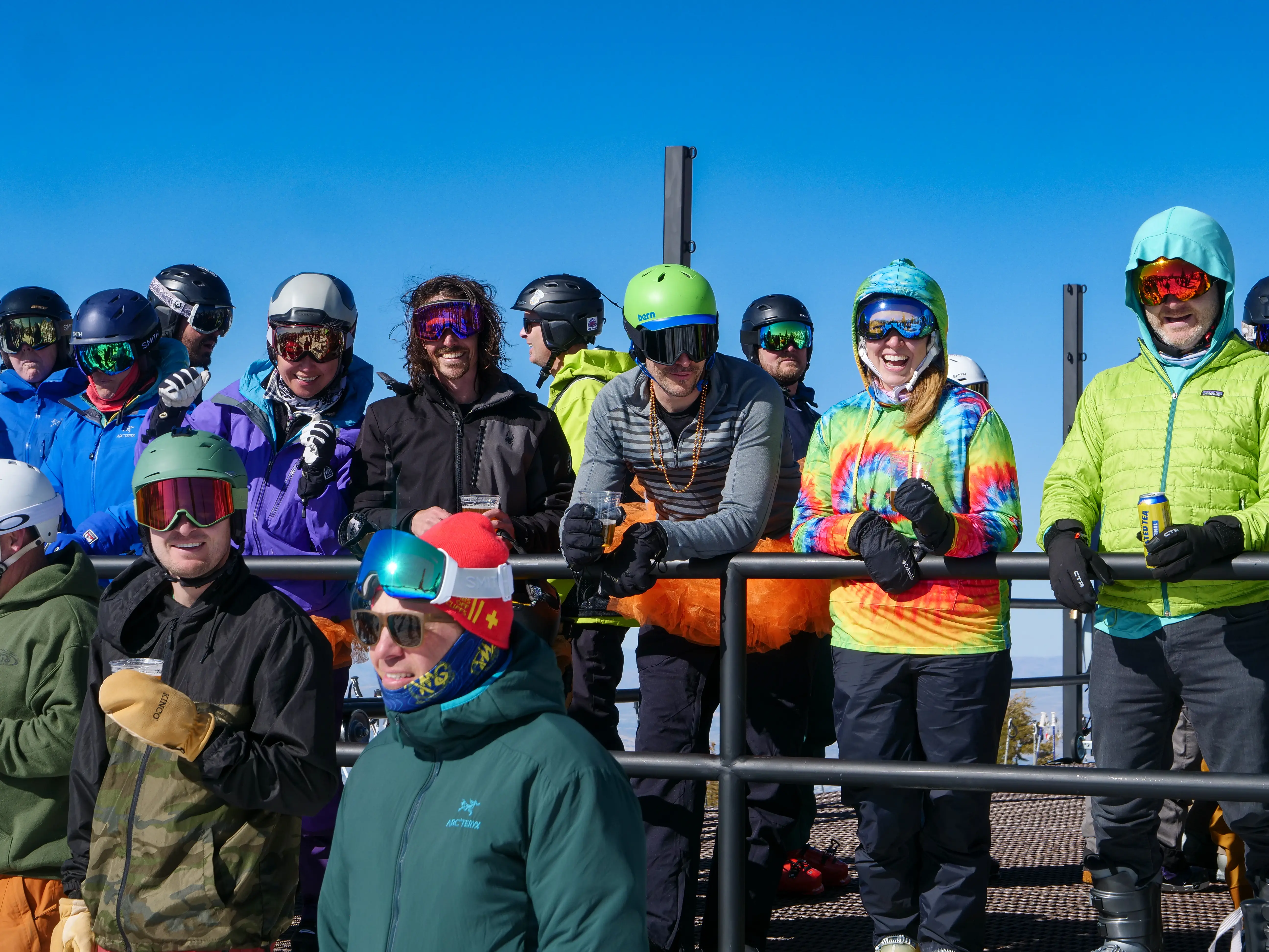 A group of skiers and snowboarders wearing colorful helmets, jackets, and goggles stand together outside on a sunny day, smiling and posing behind a metal railing with a clear blue sky in the background. Mt. Rose Ski Tahoe