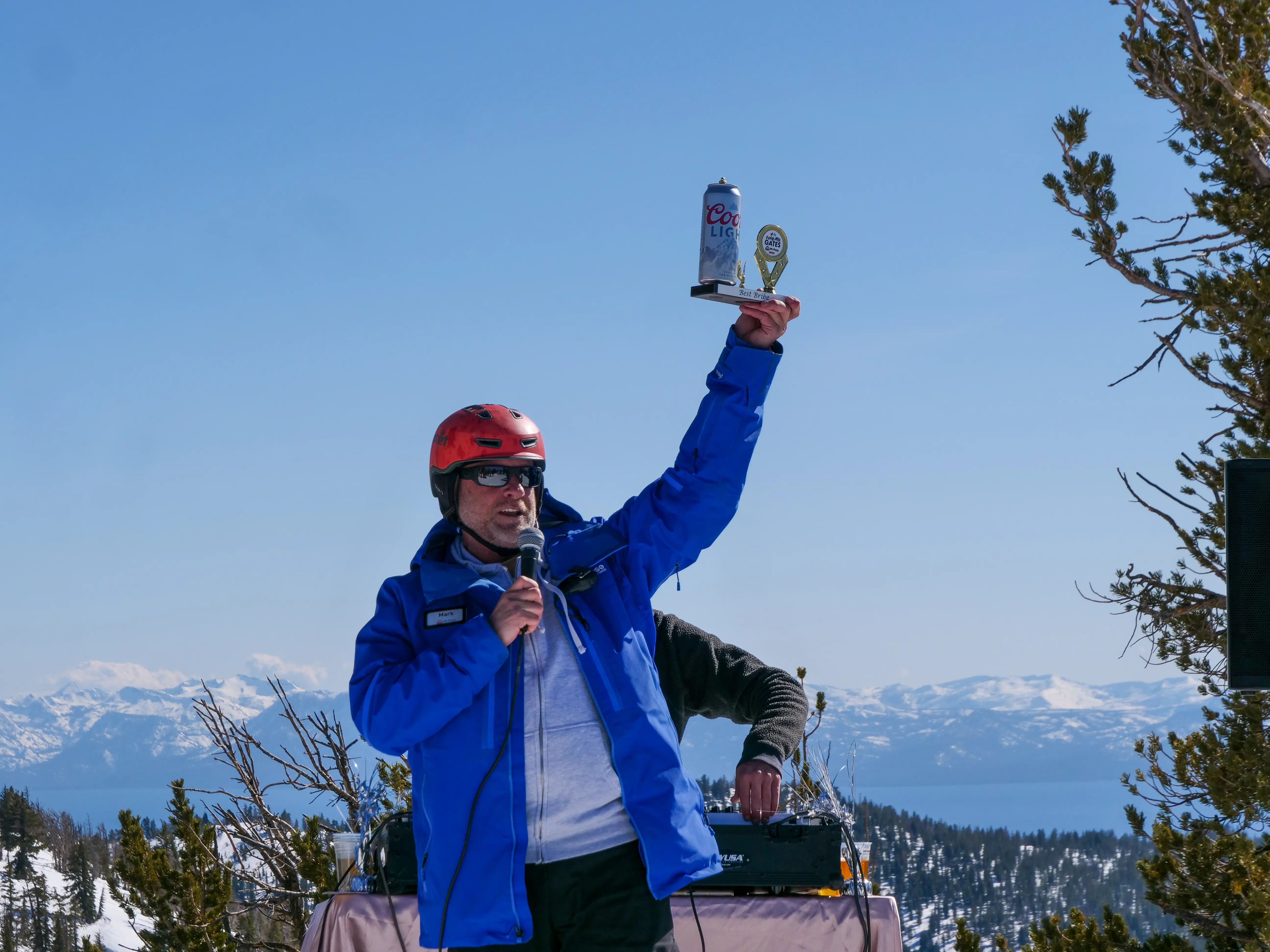 A person in a red helmet and blue jacket holds up a can of Coors Light and a small trophy while speaking into a microphone, with snowy mountains and trees in the background. Mt. Rose Ski Tahoe