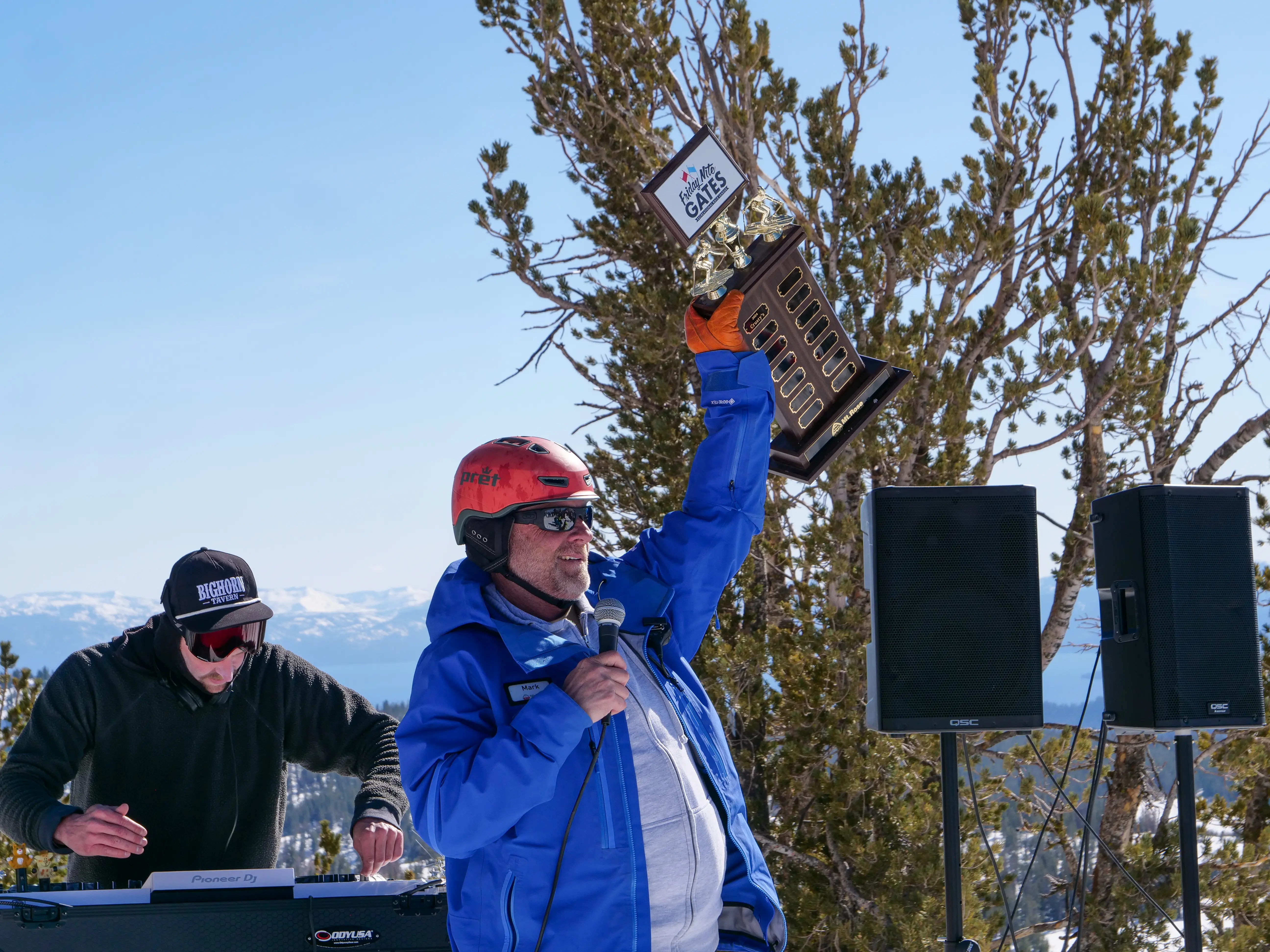 A man in a blue jacket and red helmet holds up a large trophy while speaking into a microphone outdoors, with another person wearing sunglasses and headphones standing at a DJ booth. Snow-covered mountains are visible in the background. Mt. Rose Ski Tahoe