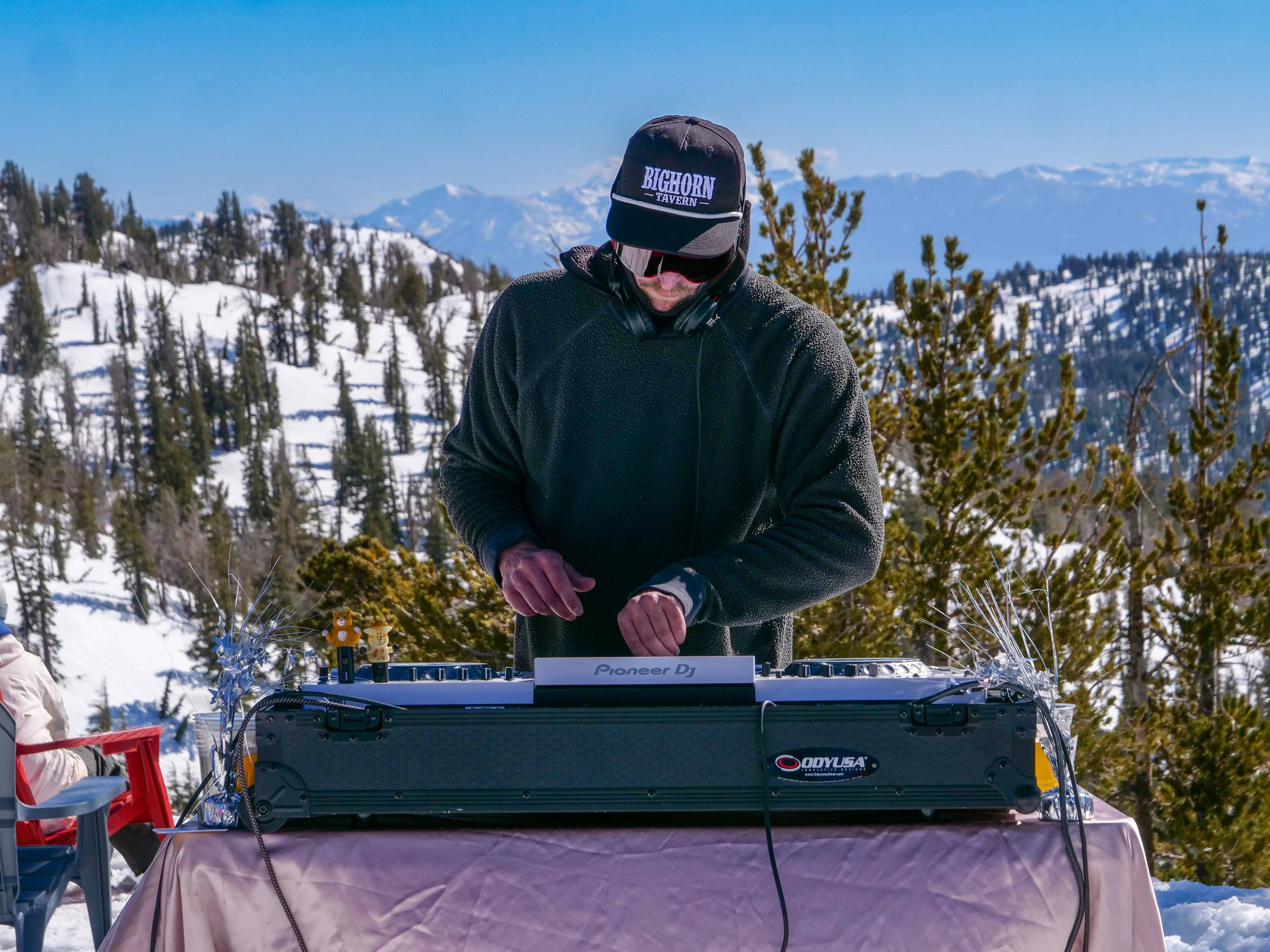 A DJ wearing sunglasses and a cap is playing music on a Pioneer DJ controller outdoors, with snowy mountains and pine trees in the background. Mt. Rose Ski Tahoe