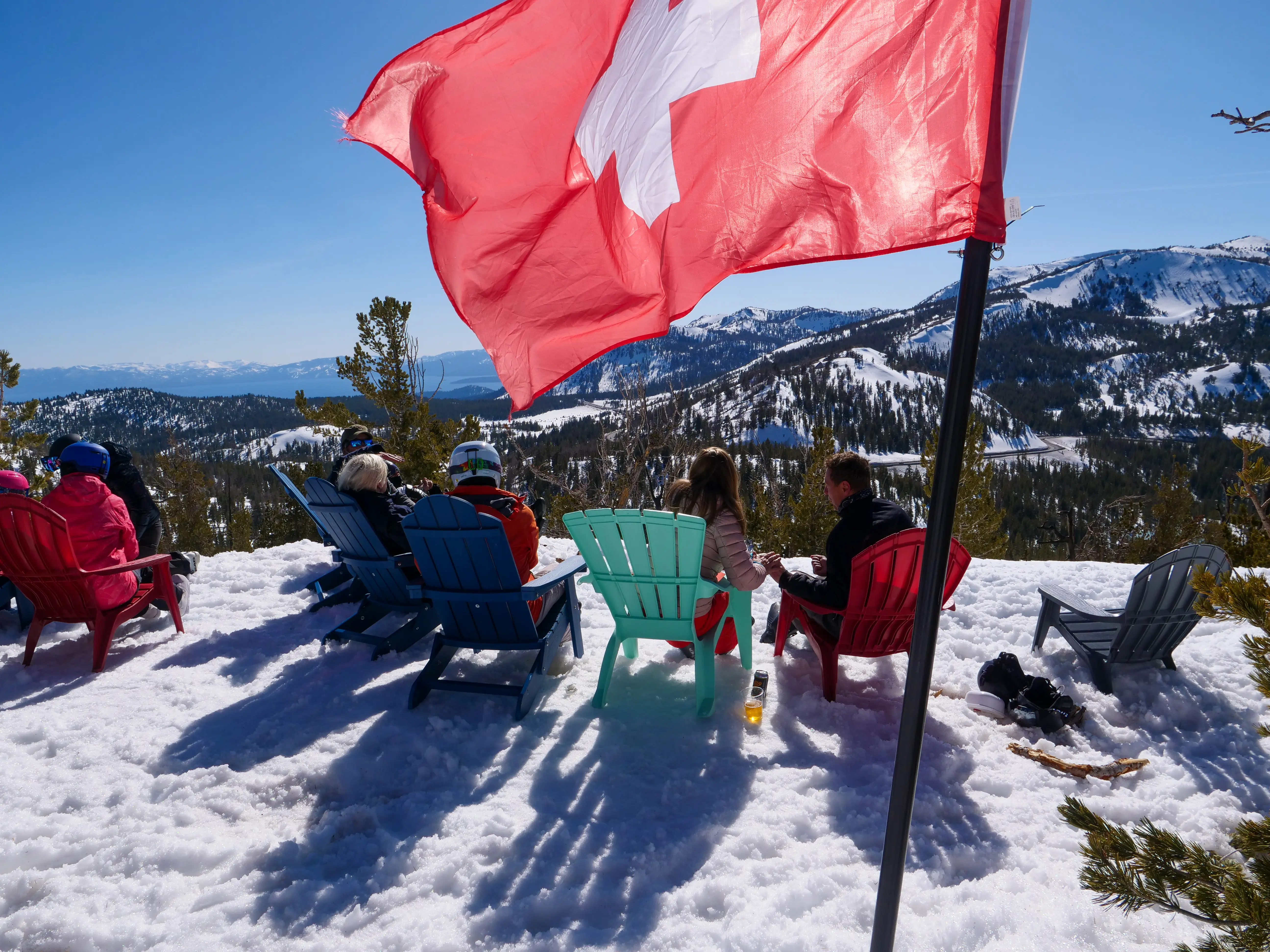 People sit in colorful chairs on a snowy mountain slope, enjoying the view of surrounding peaks. A red Swiss flag waves in the foreground under a clear blue sky. Mt. Rose Ski Tahoe