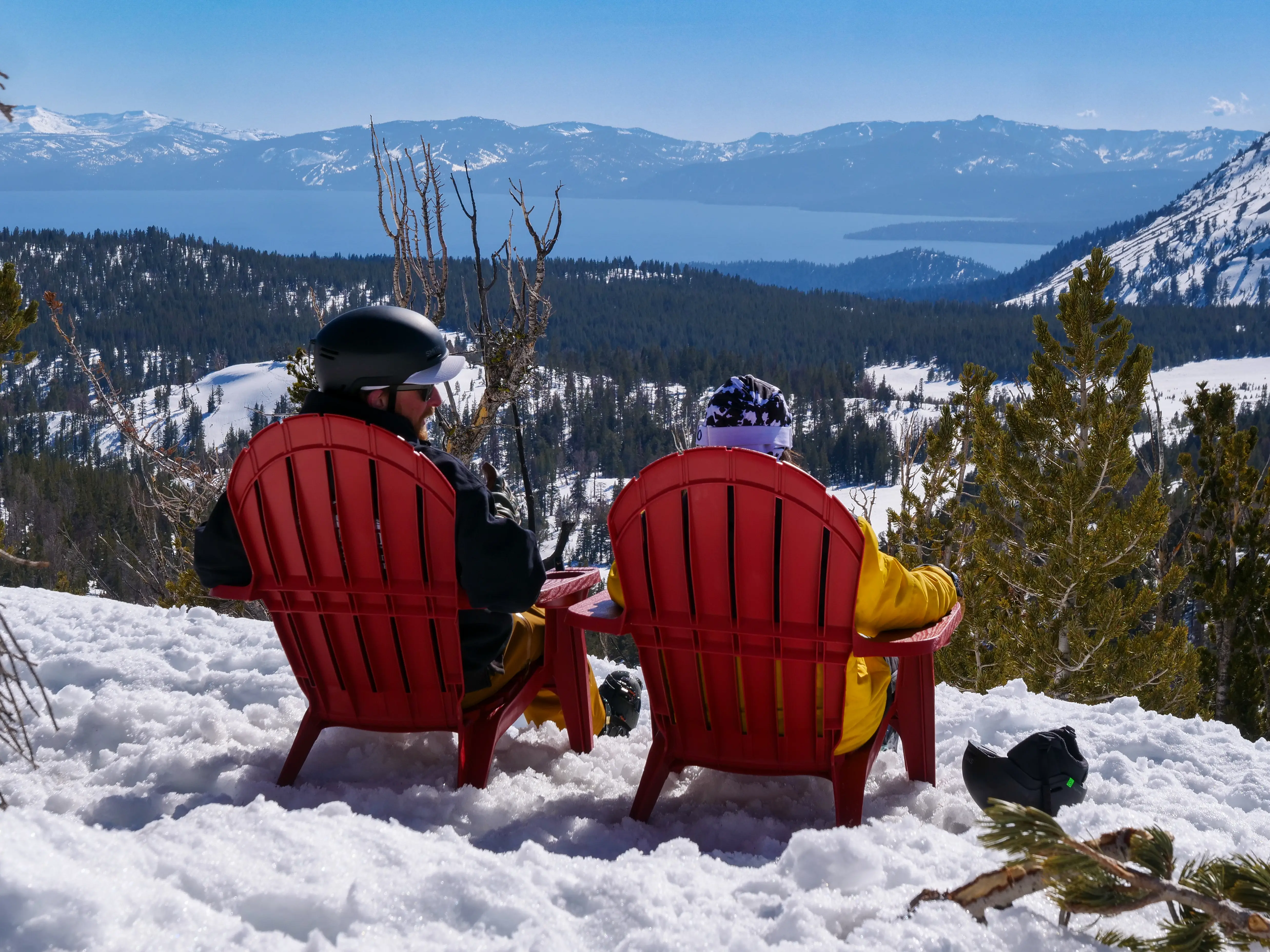 Two people in winter gear sit in red Adirondack chairs on snow, overlooking a forested mountain landscape and a large lake under a clear blue sky. Mt. Rose Ski Tahoe