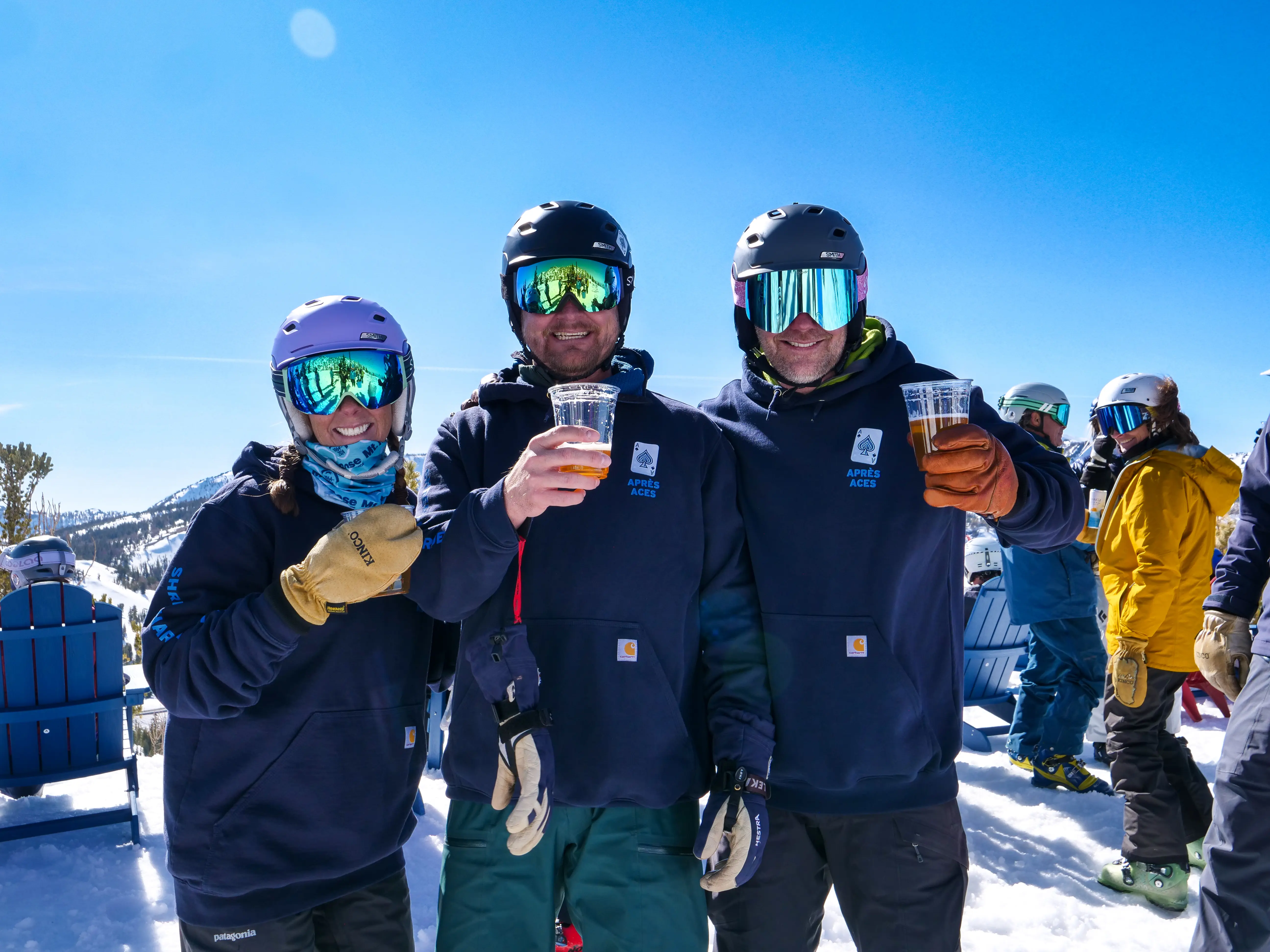 Three people in ski gear and helmets, smiling and holding drinks, stand together on a snowy mountain with other skiers and bright blue sky in the background. Mt. Rose Ski Tahoe