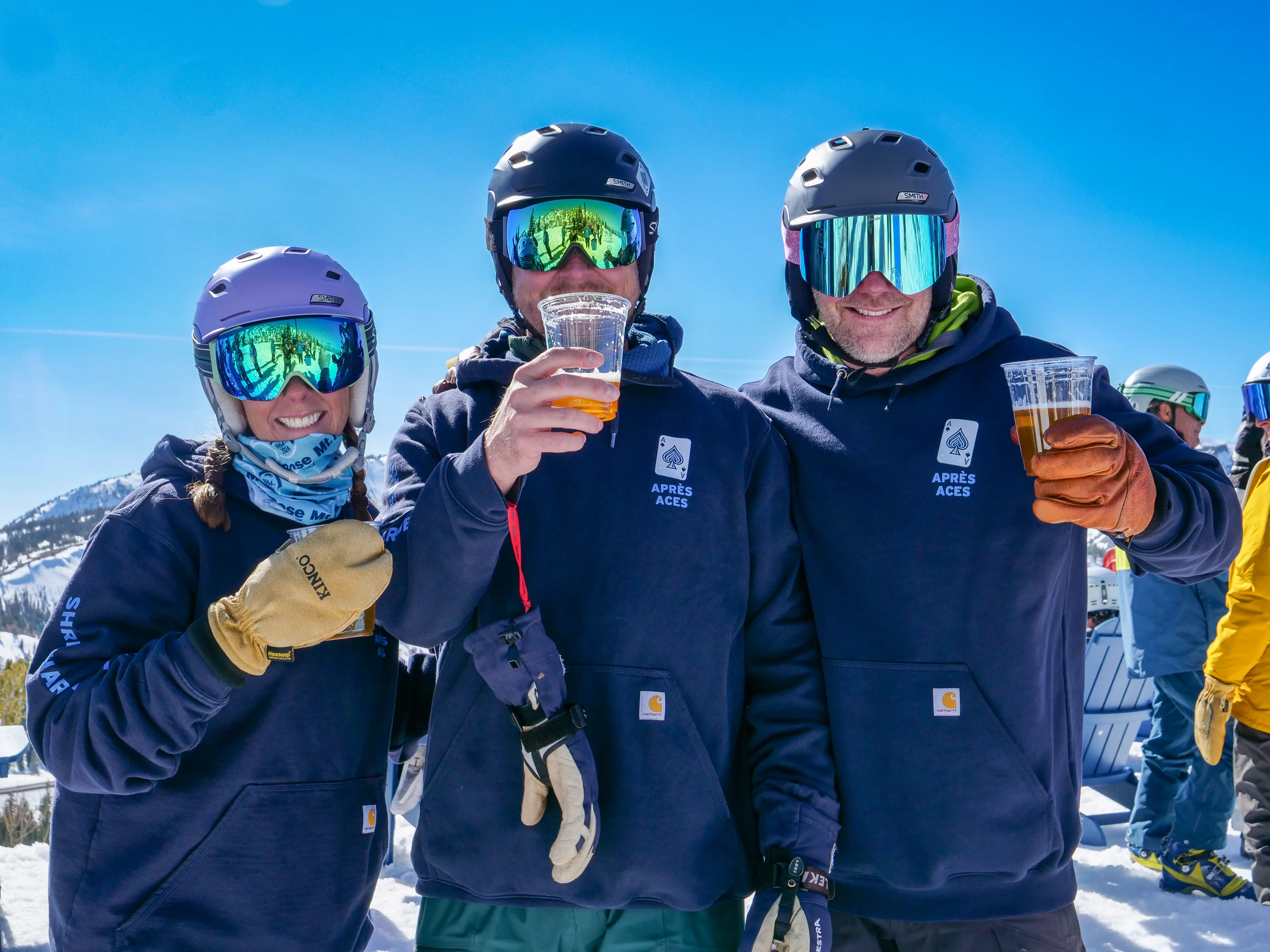 Three people in ski gear and blue hoodies smile and hold drinks while standing outdoors in the snow on a sunny day, with snowy mountains and other skiers in the background. Mt. Rose Ski Tahoe
