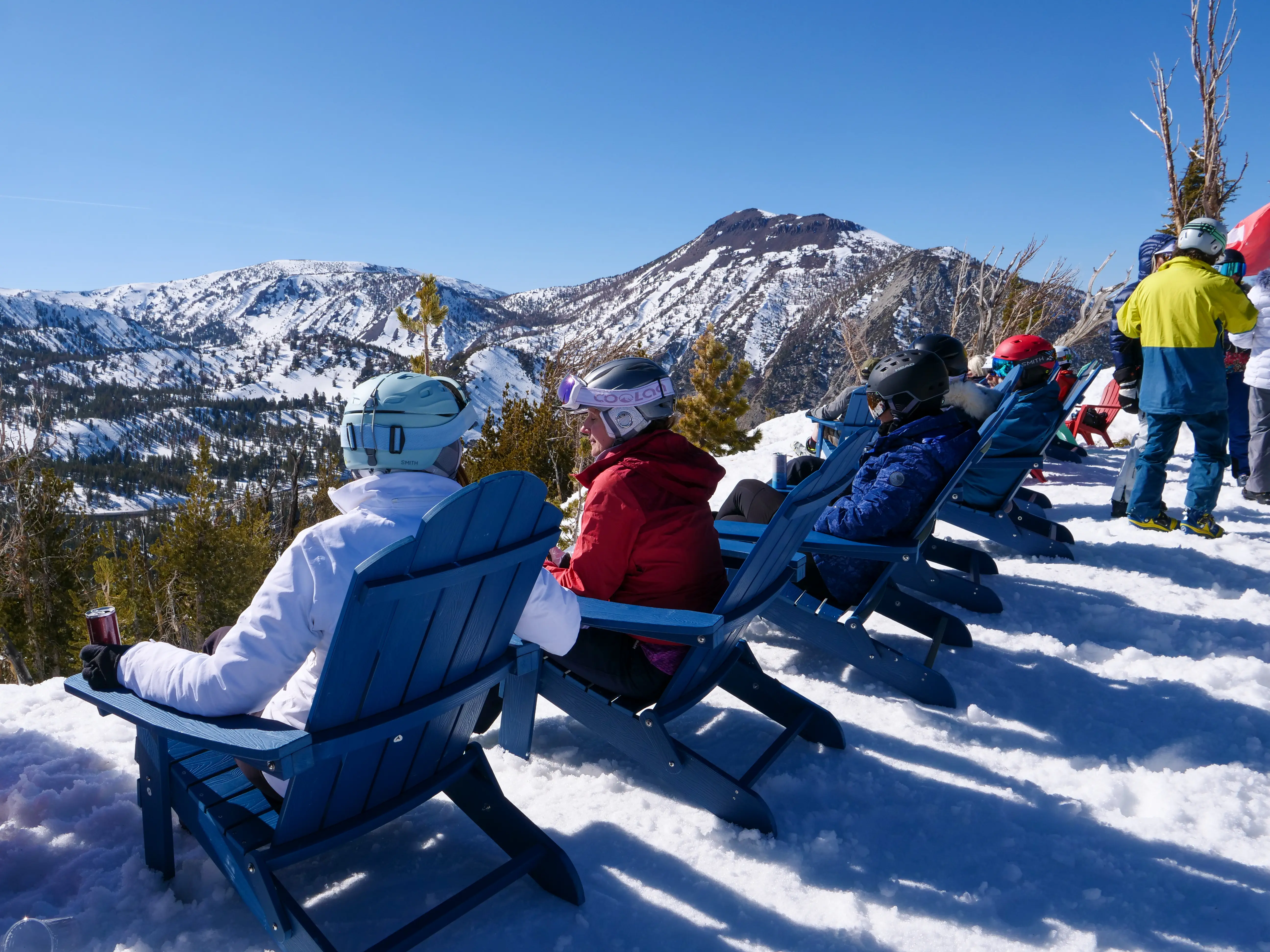 People in ski gear sit on blue Adirondack chairs in the snow, enjoying a scenic mountain view under a clear blue sky. Snow-covered peaks and pine trees are visible in the background. Mt. Rose Ski Tahoe