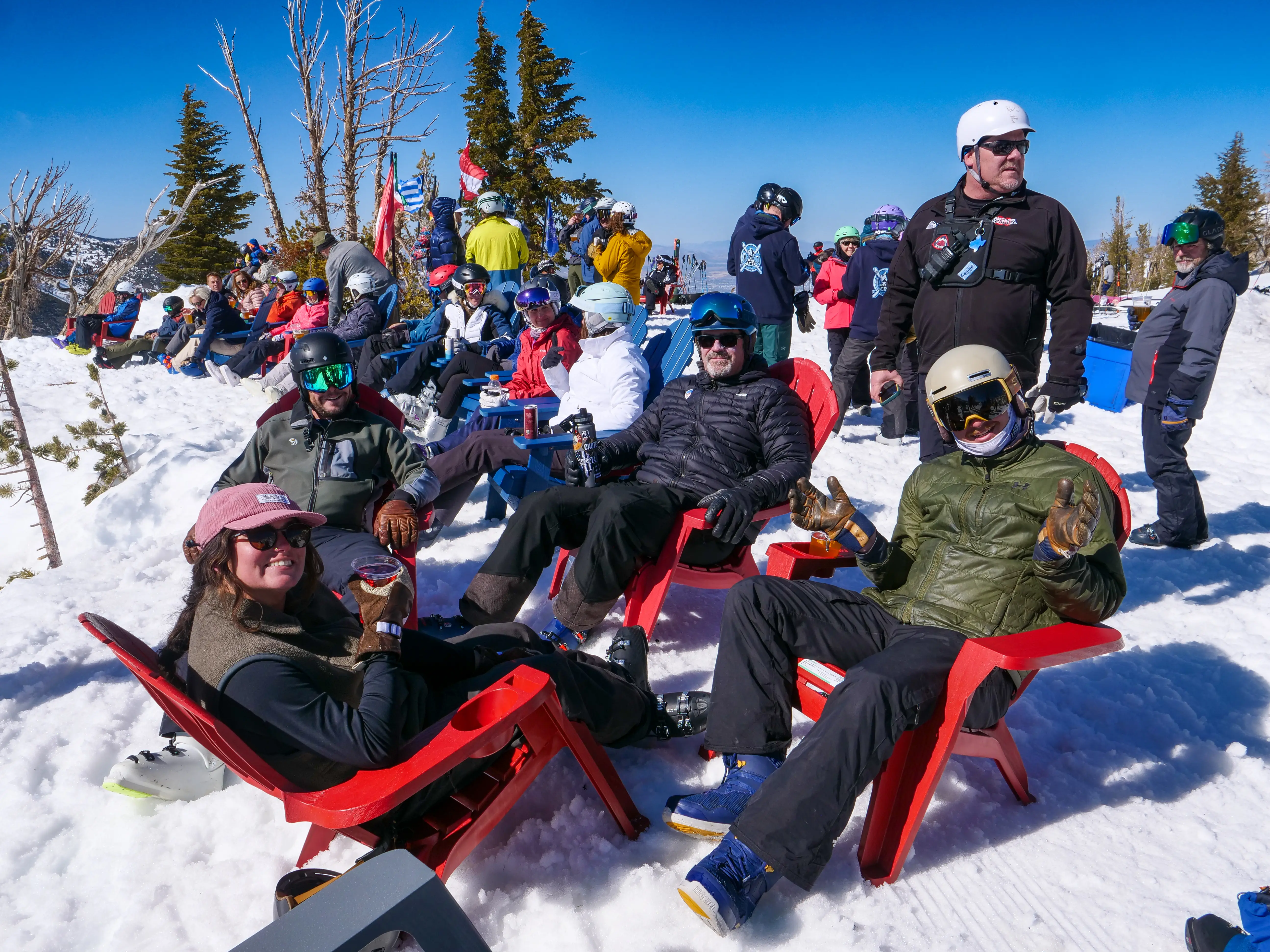 A group of people in winter gear relax in red chairs on snowy ground, enjoying drinks, with skiers and snowboarders gathered in the background under a clear blue sky. Mt. Rose Ski Tahoe