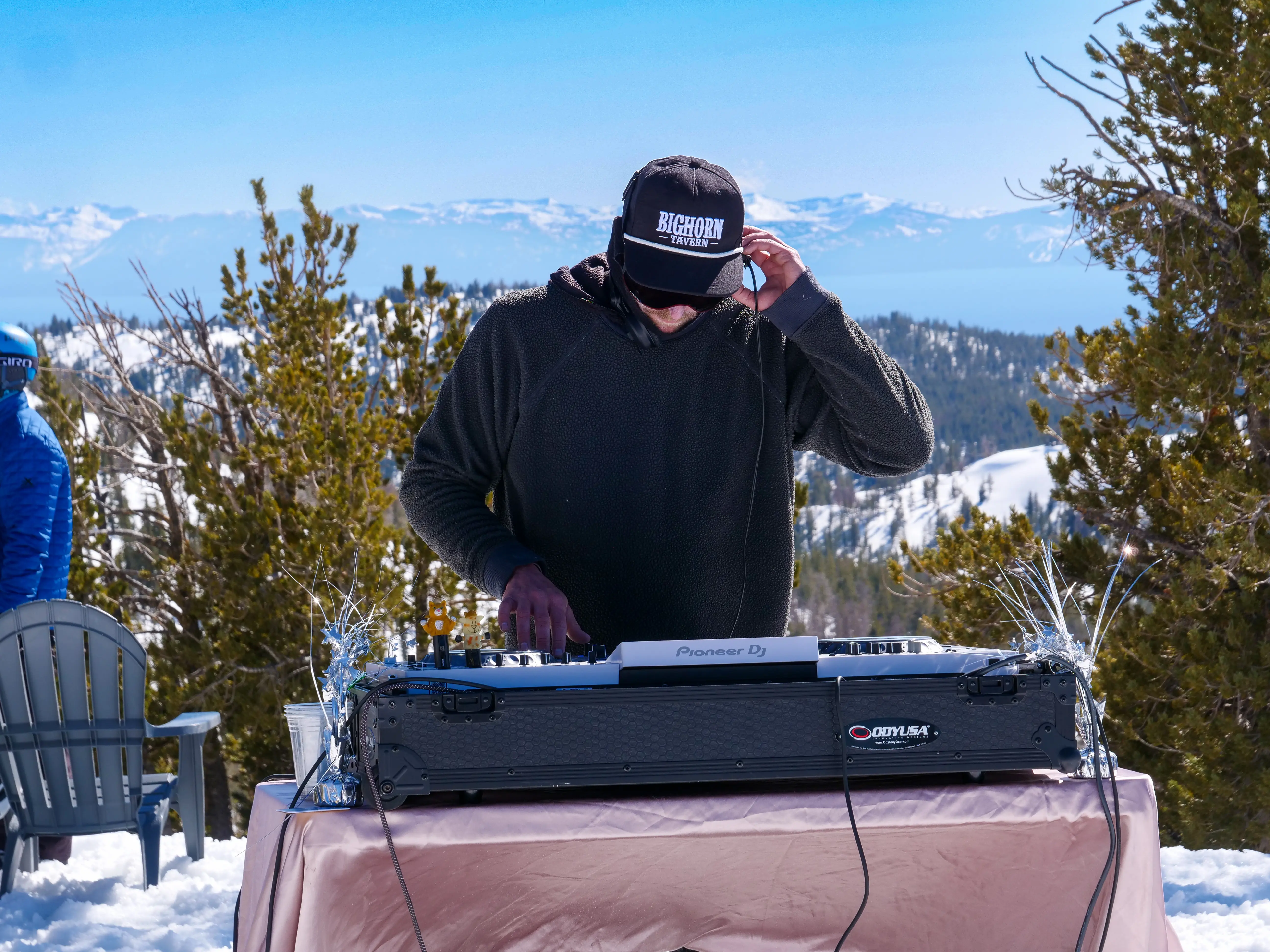 A DJ wearing a cap and sweater performs outdoors on snowy mountains, using a Pioneer DJ controller placed on a table covered with a pink cloth, with pine trees and a scenic view in the background. Mt. Rose Ski Tahoe