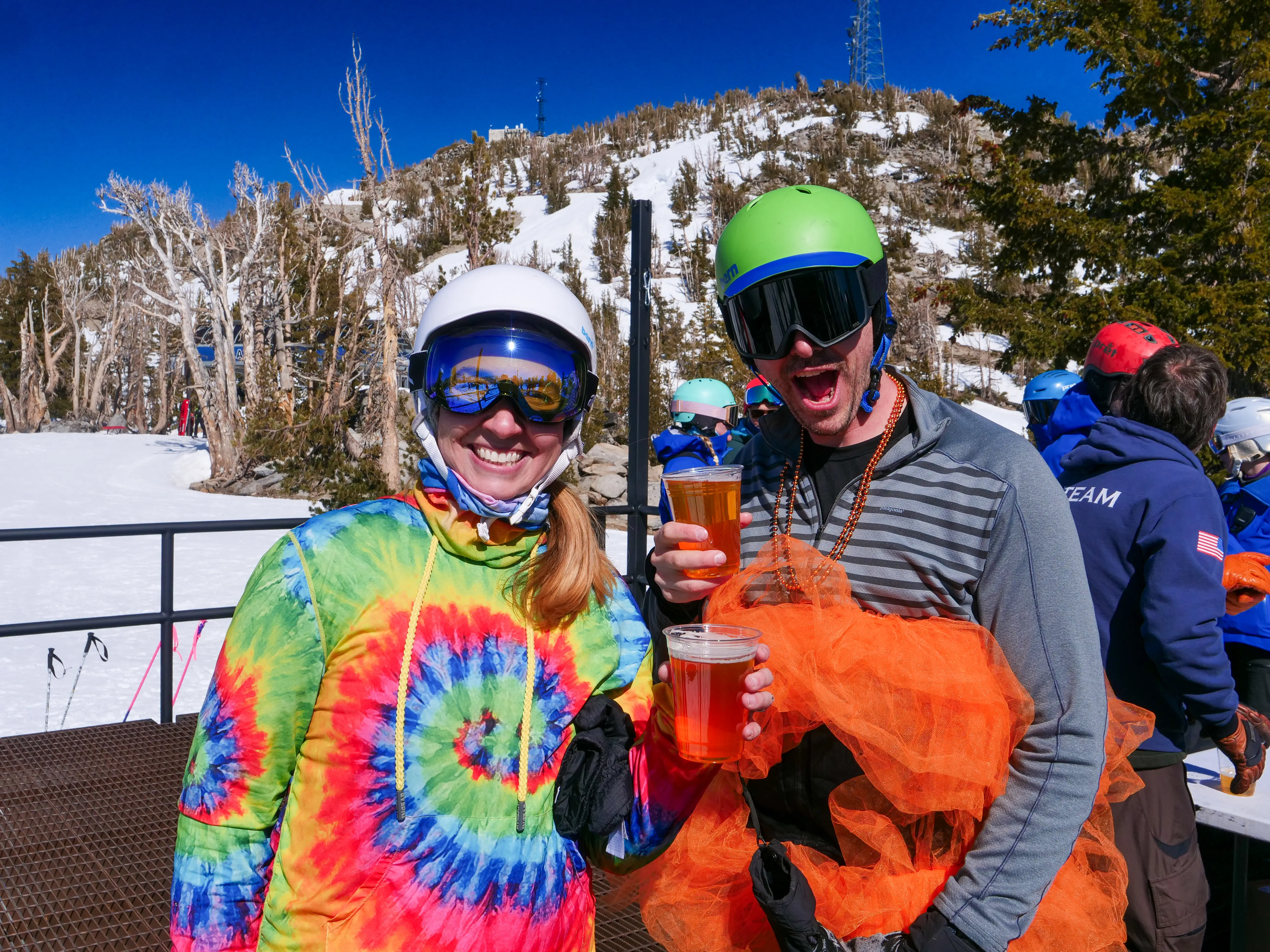 Two skiers in colorful outfits and helmets smile while holding cups of beer on a snowy mountain with people and trees in the background under a clear blue sky. Mt. Rose Ski Tahoe