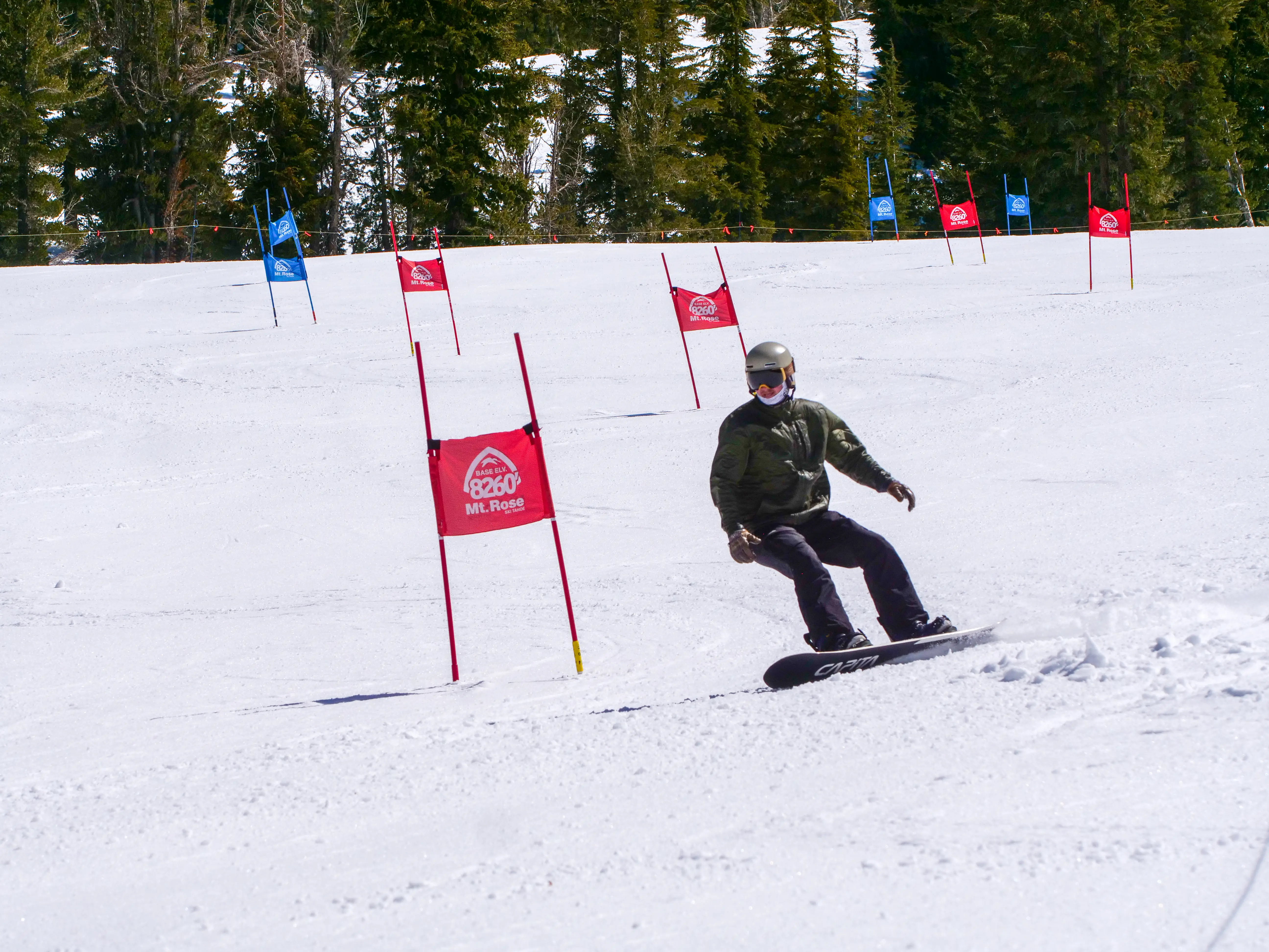 A snowboarder in a green jacket and helmet rides down a snowy slope, maneuvering through red and blue slalom gates on a sunny day with green trees in the background. Mt. Rose Ski Tahoe