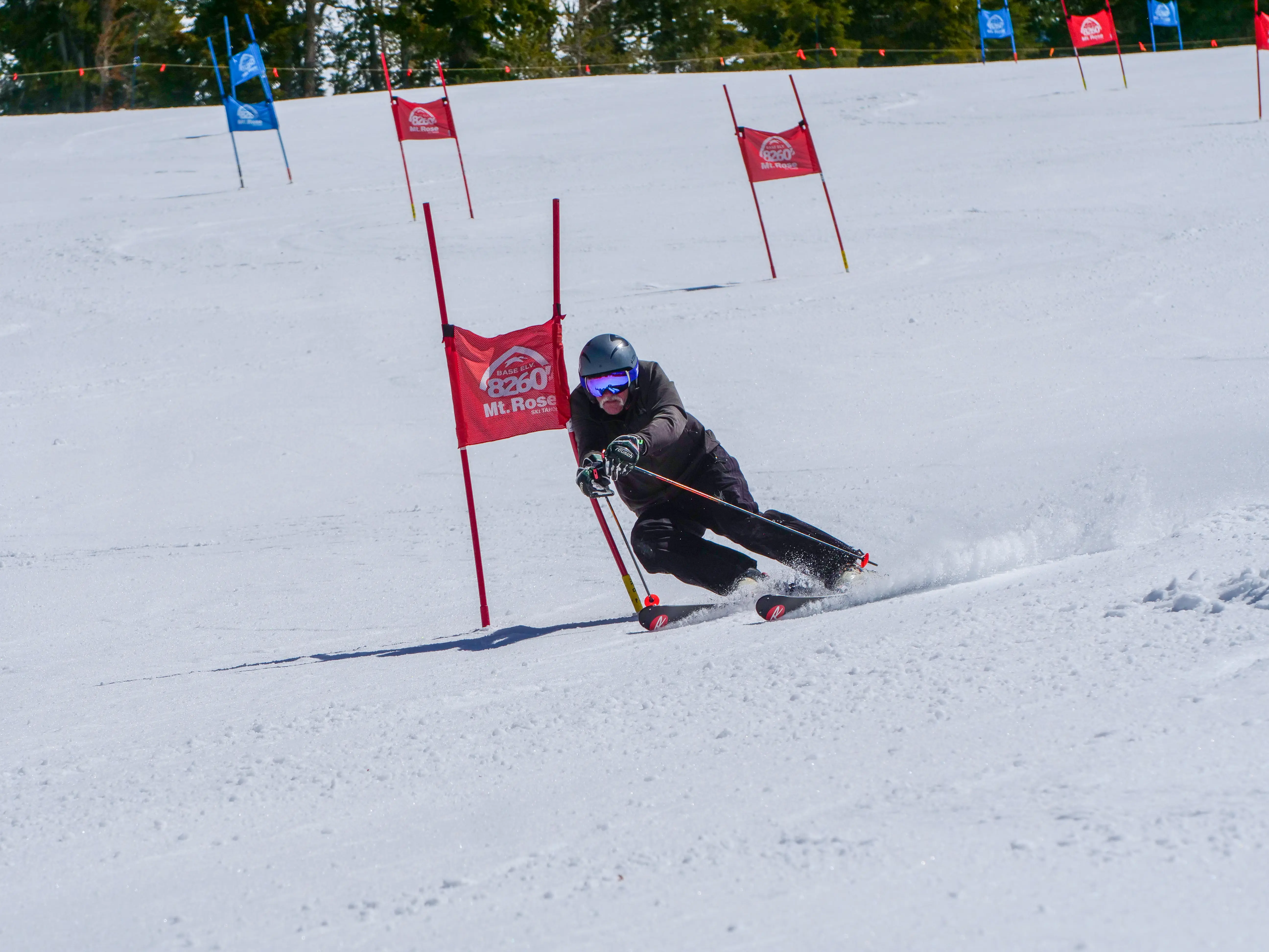A skier in black gear and helmet makes a sharp turn around a red slalom gate on a snowy mountain race course, with more gates visible in the background. Mt. Rose Ski Tahoe