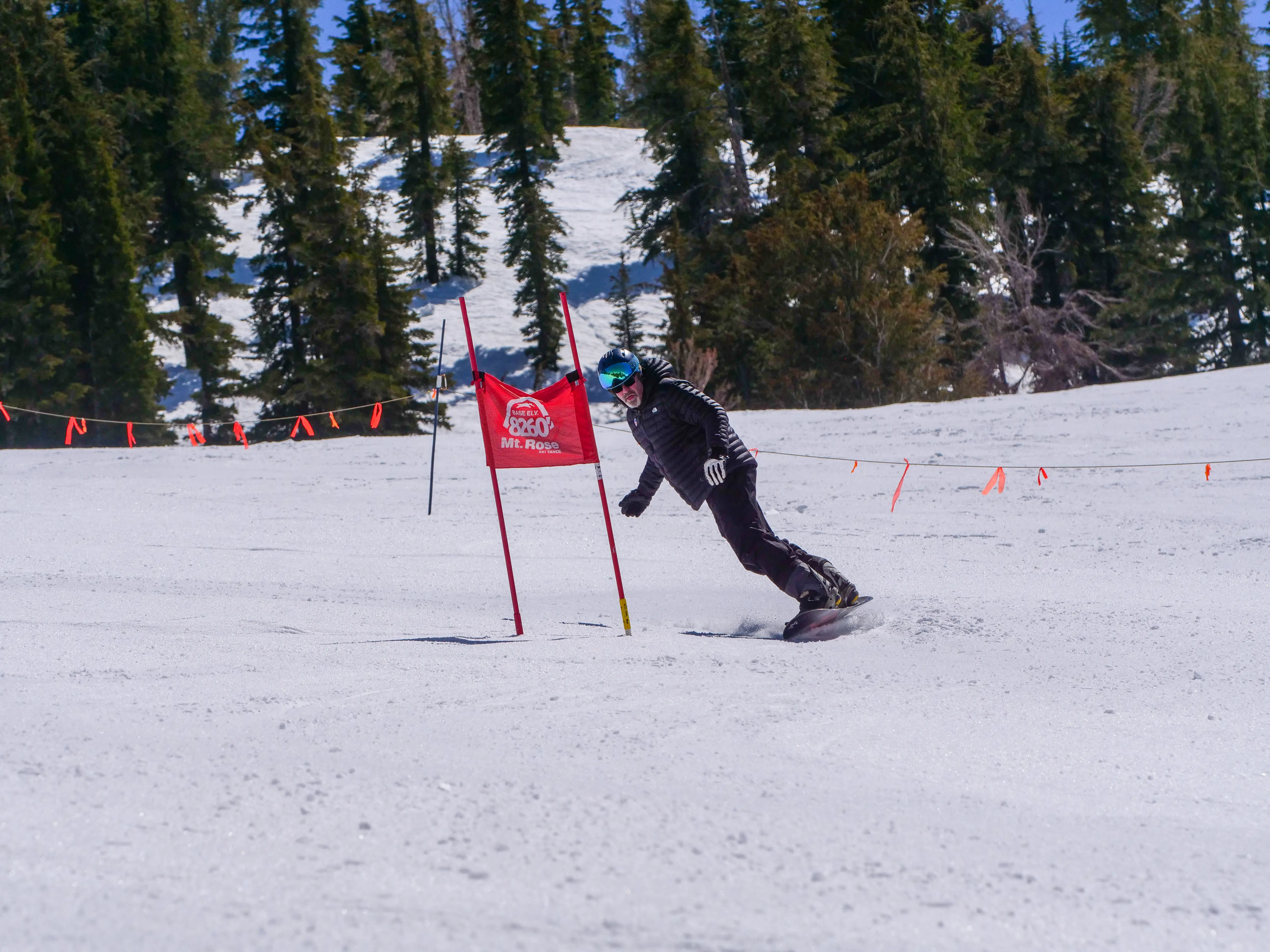 A snowboarder in a black outfit and blue helmet is racing downhill around a red slalom gate marked "Mt. Rose" on a snowy slope, with pine trees and blue sky in the background. Mt. Rose Ski Tahoe