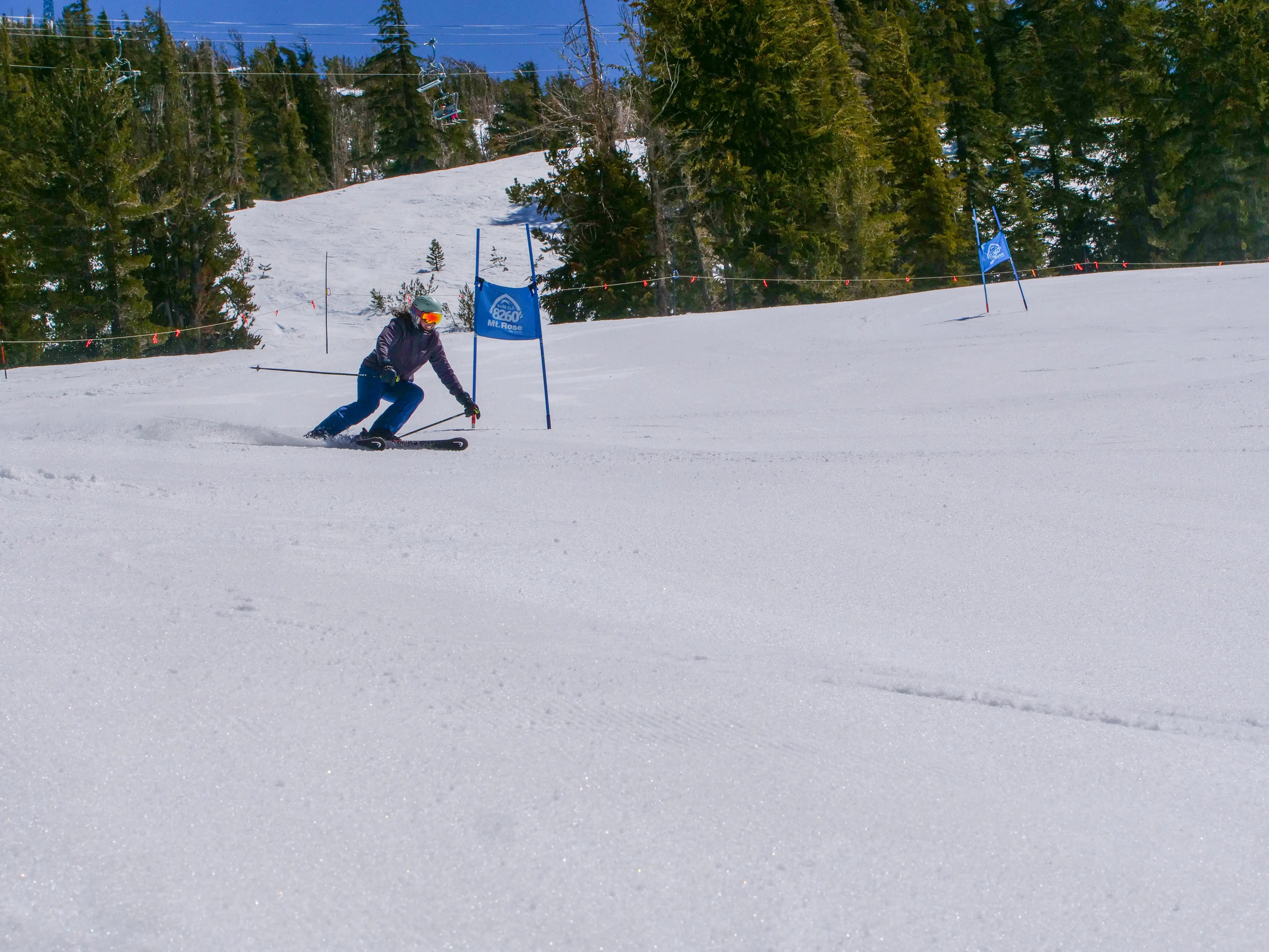 A skier wearing a helmet and goggles makes a sharp turn on a snowy slope during a slalom race, passing blue marker flags, with evergreen trees and ski lifts visible in the background under a clear sky. Mt. Rose Ski Tahoe