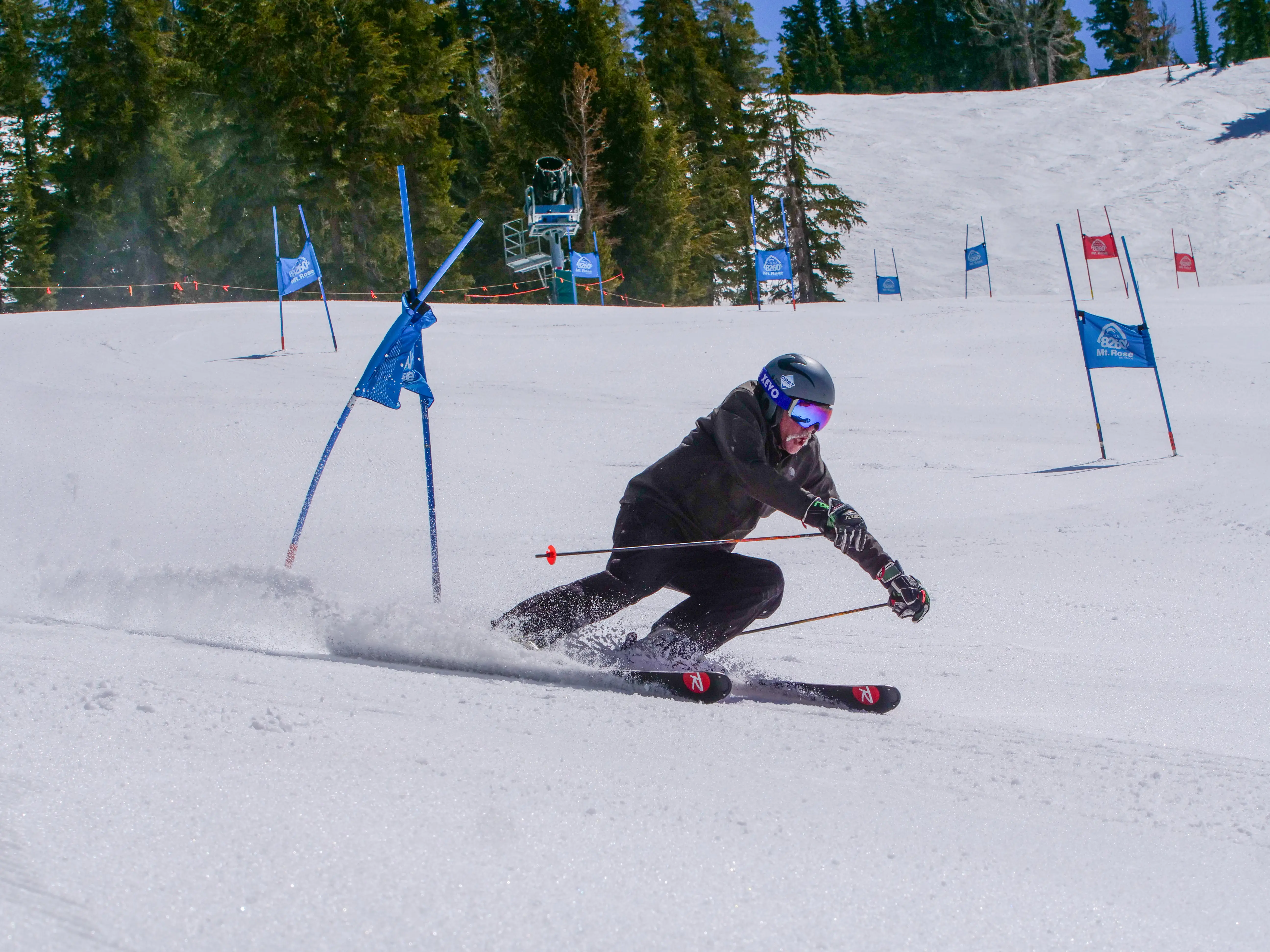 A skier in black gear and a helmet makes a sharp turn around a blue slalom gate on a snowy mountain slope, surrounded by pine trees and blue flags under a clear sky. Mt. Rose Ski Tahoe