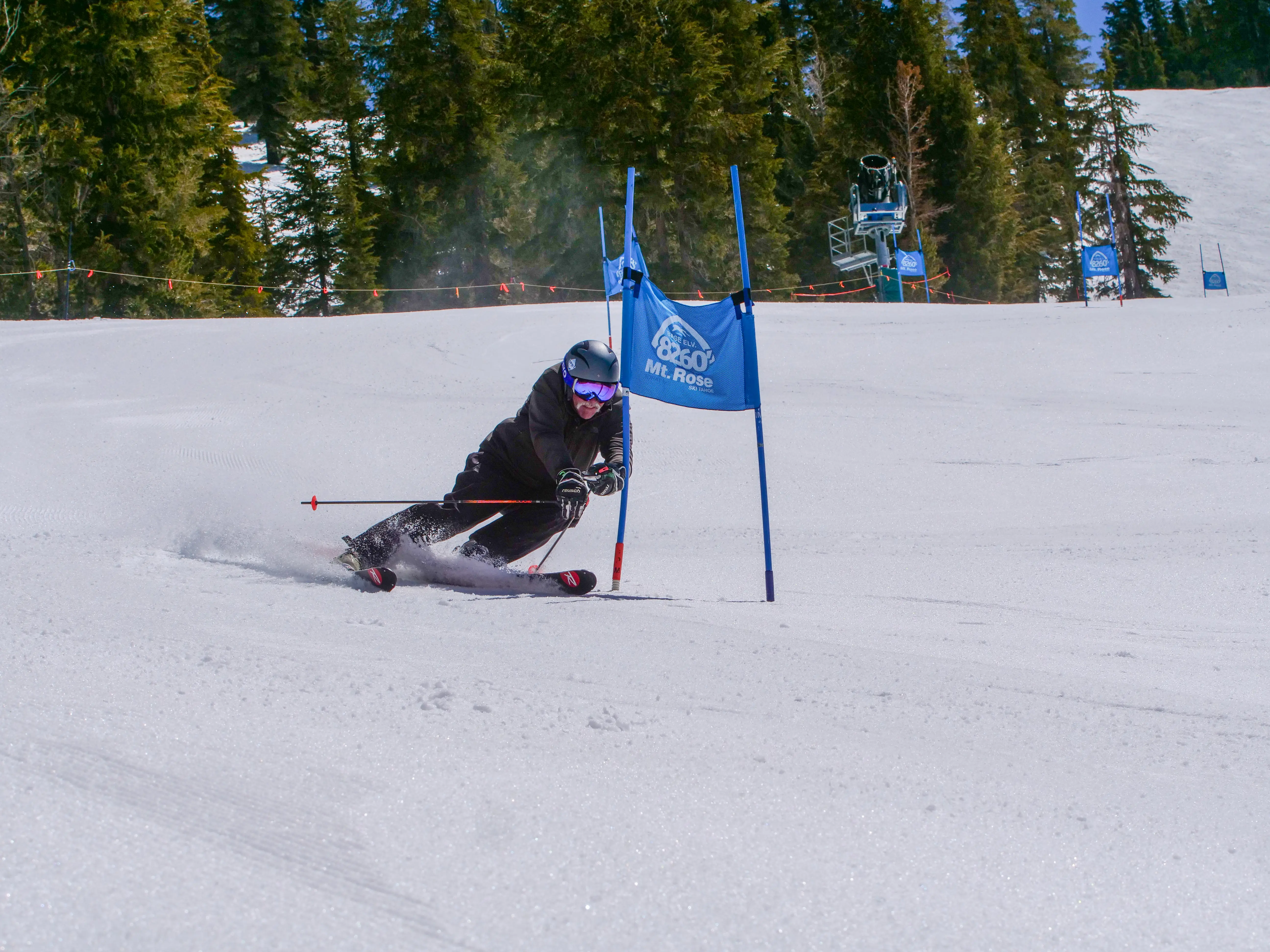 A skier wearing a helmet and goggles leans sharply while navigating a slalom gate on a snowy slope at Mt. Rose, surrounded by trees under a clear sky. Mt. Rose Ski Tahoe