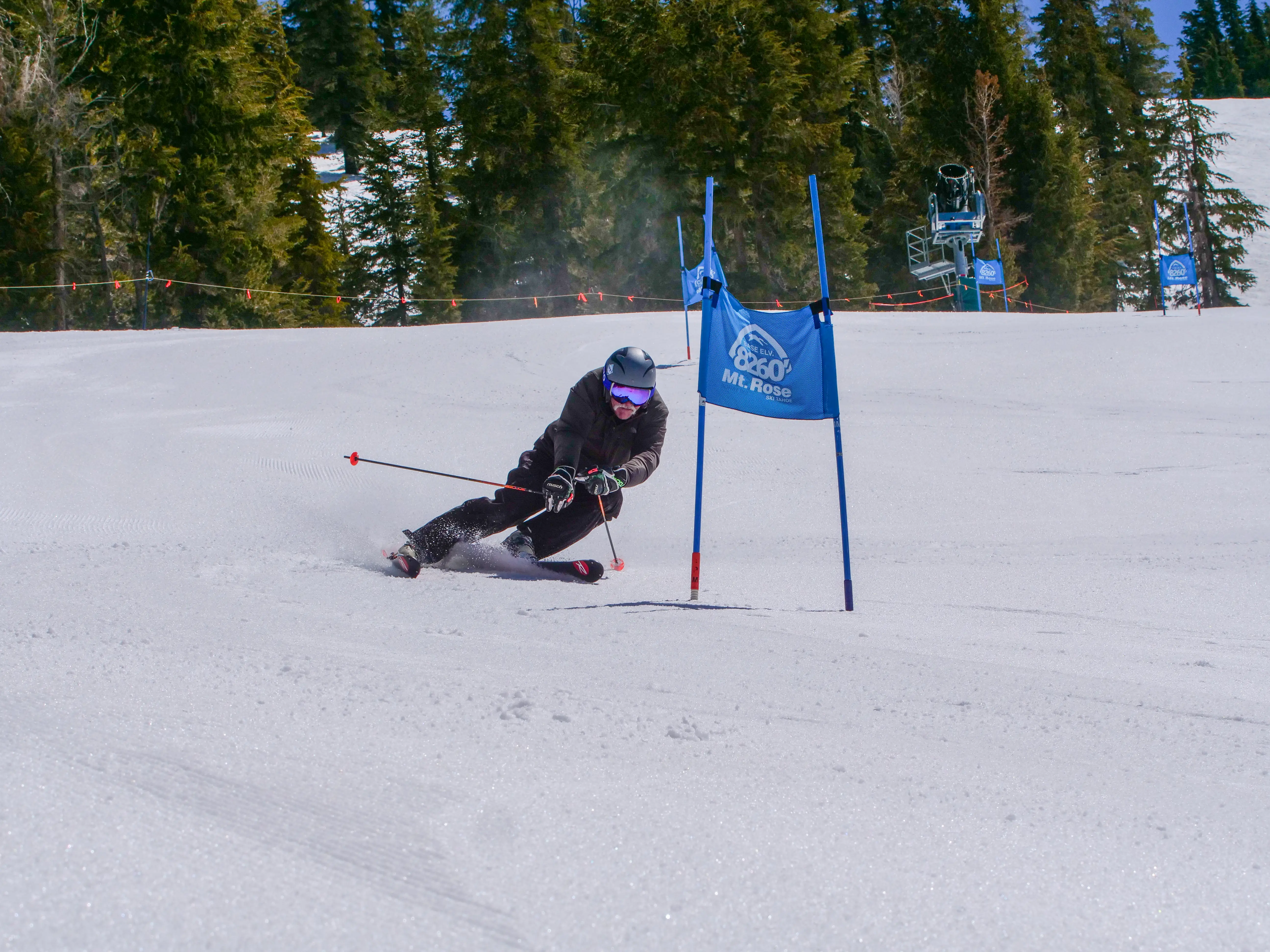 A skier in black gear and a helmet races downhill, leaning sharply around a blue gate labeled "Mt. Rose," on a snowy slope with pine trees and ski lift in the background. Mt. Rose Ski Tahoe