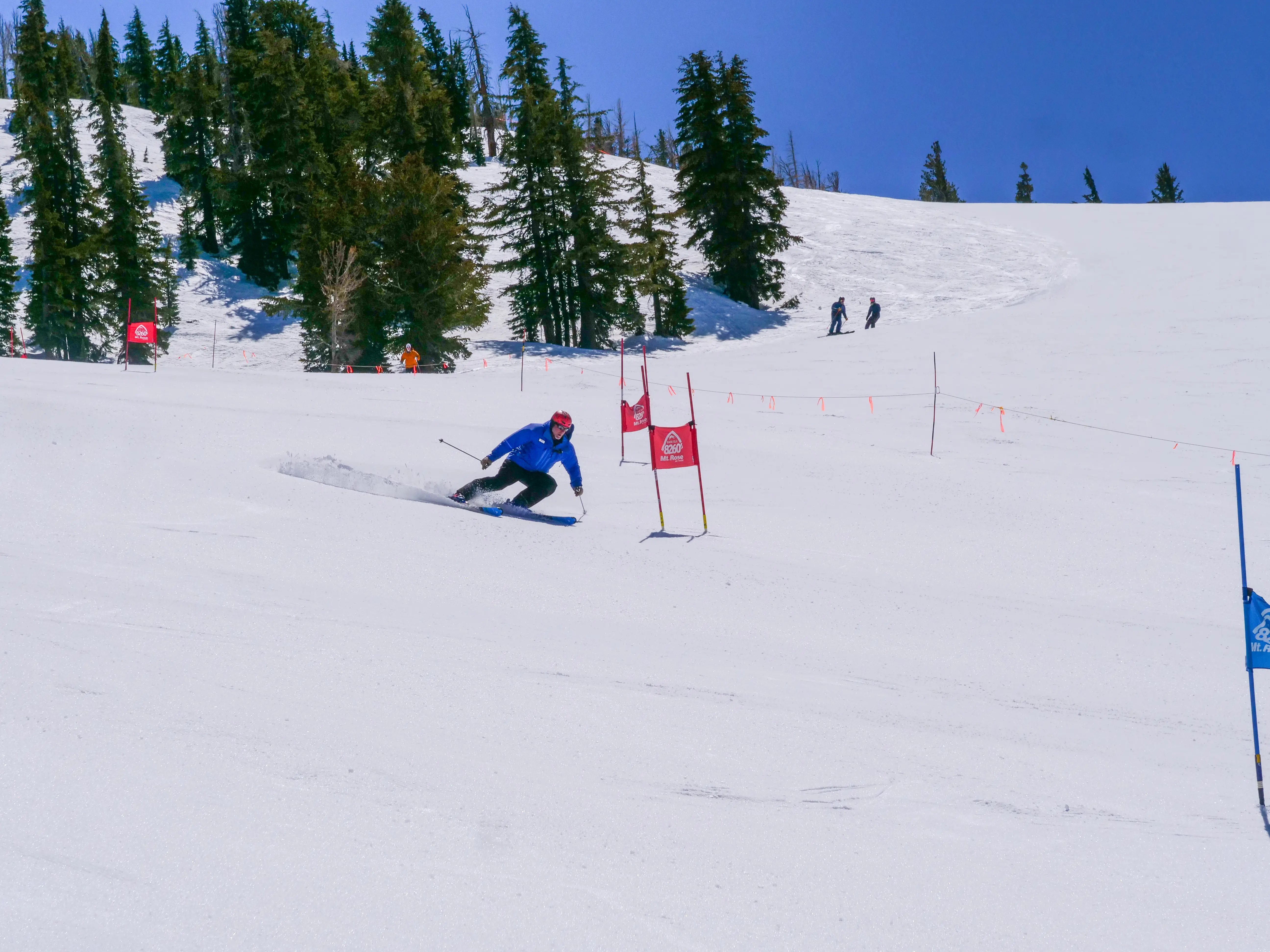 A skier in a blue jacket and red helmet races downhill between slalom gates on a snowy slope, with evergreen trees and a few people in the background under a clear blue sky. Mt. Rose Ski Tahoe