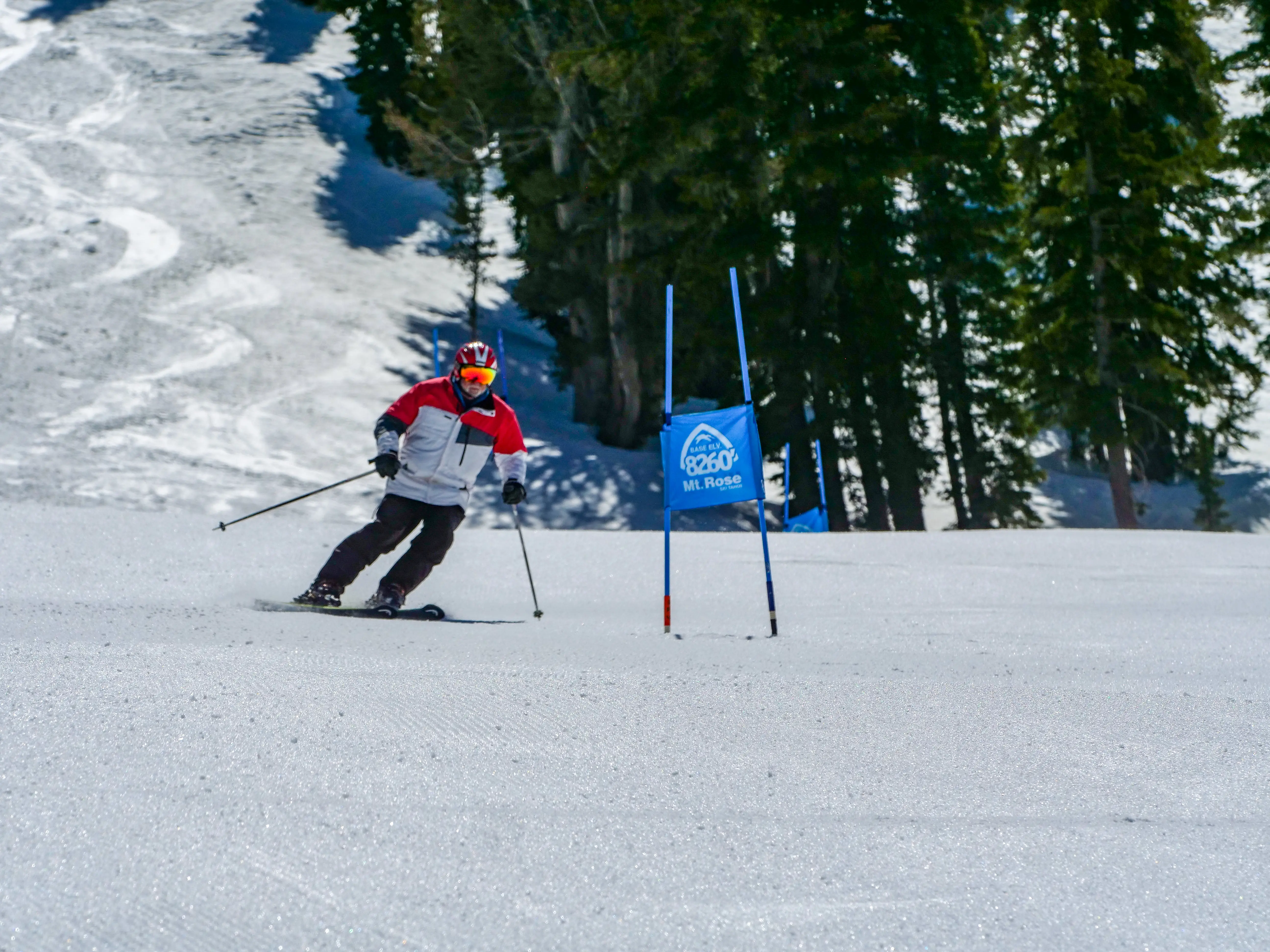 A skier wearing a red and white jacket and helmet navigates a slalom course on a snowy slope near a blue gate sign labeled "Mt. Reba," with pine trees visible in the background. Mt. Rose Ski Tahoe