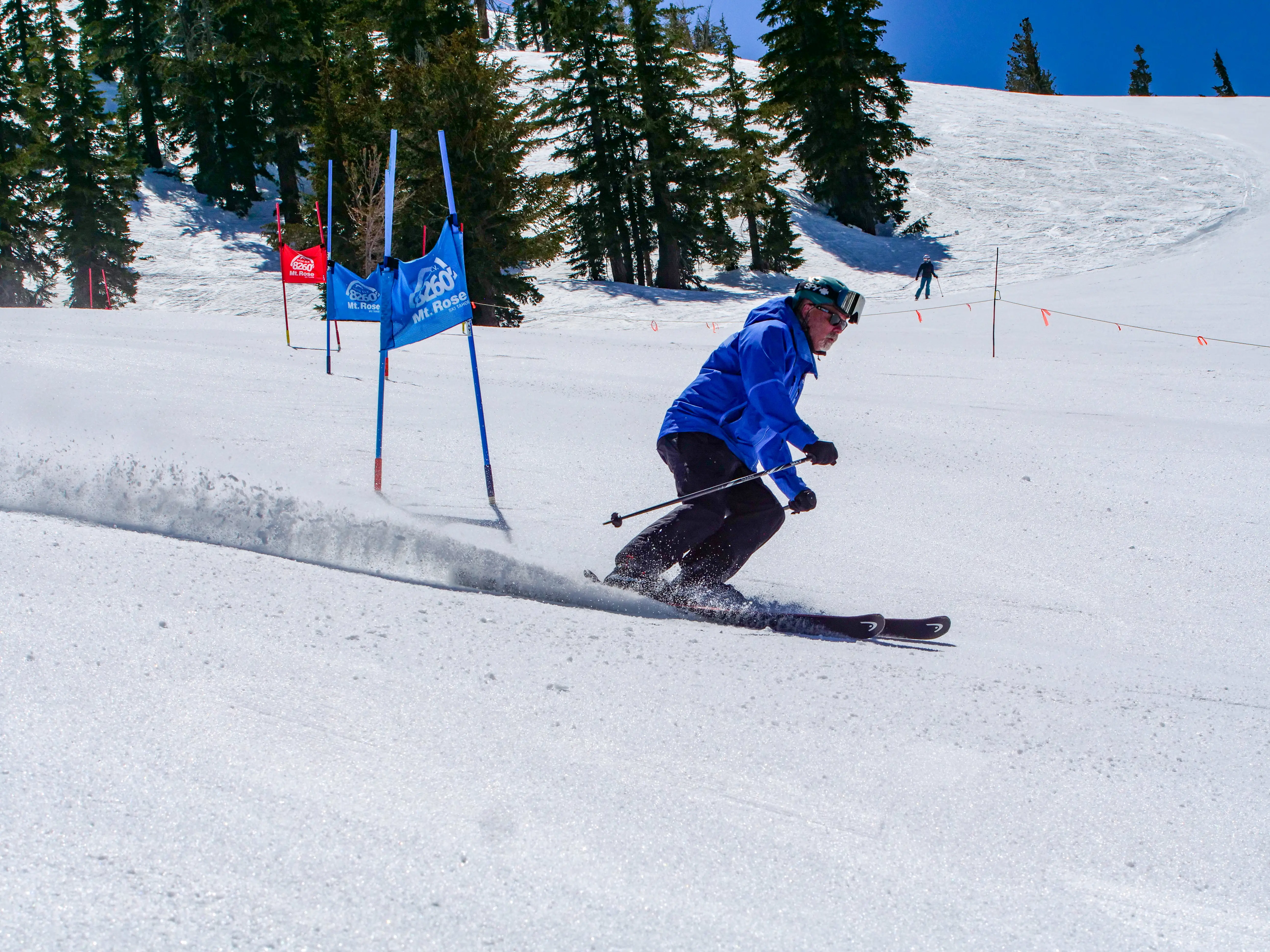 A skier in a blue jacket and black pants navigates a downhill slalom course on a snowy mountain slope, kicking up powder, with evergreen trees and blue flags in the background. Mt. Rose Ski Tahoe