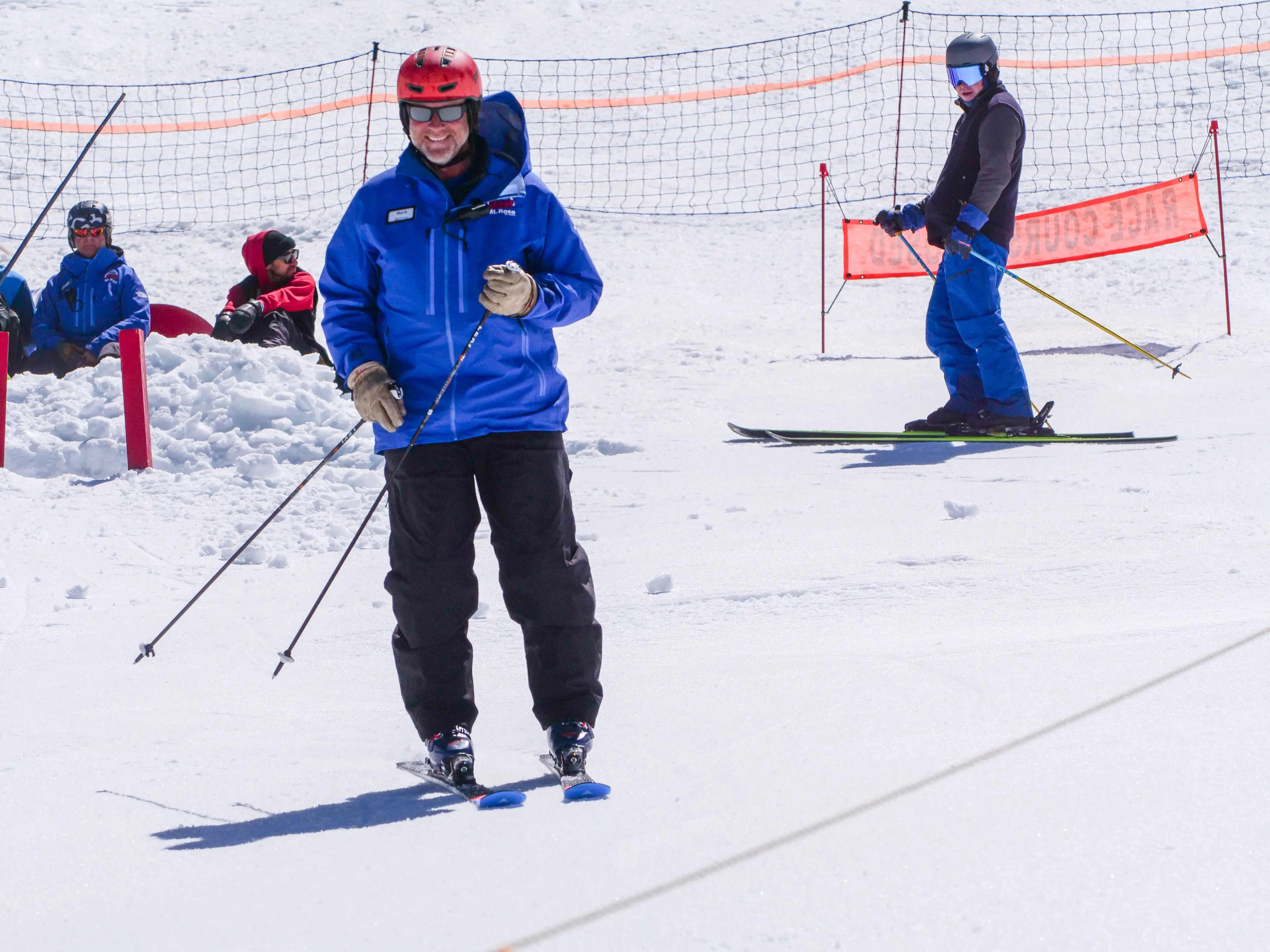 A person in a blue jacket and red helmet skis downhill, smiling. In the background, another skier stands near an orange net while two people sit on the snow. The scene is bright and snowy. Mt. Rose Ski Tahoe