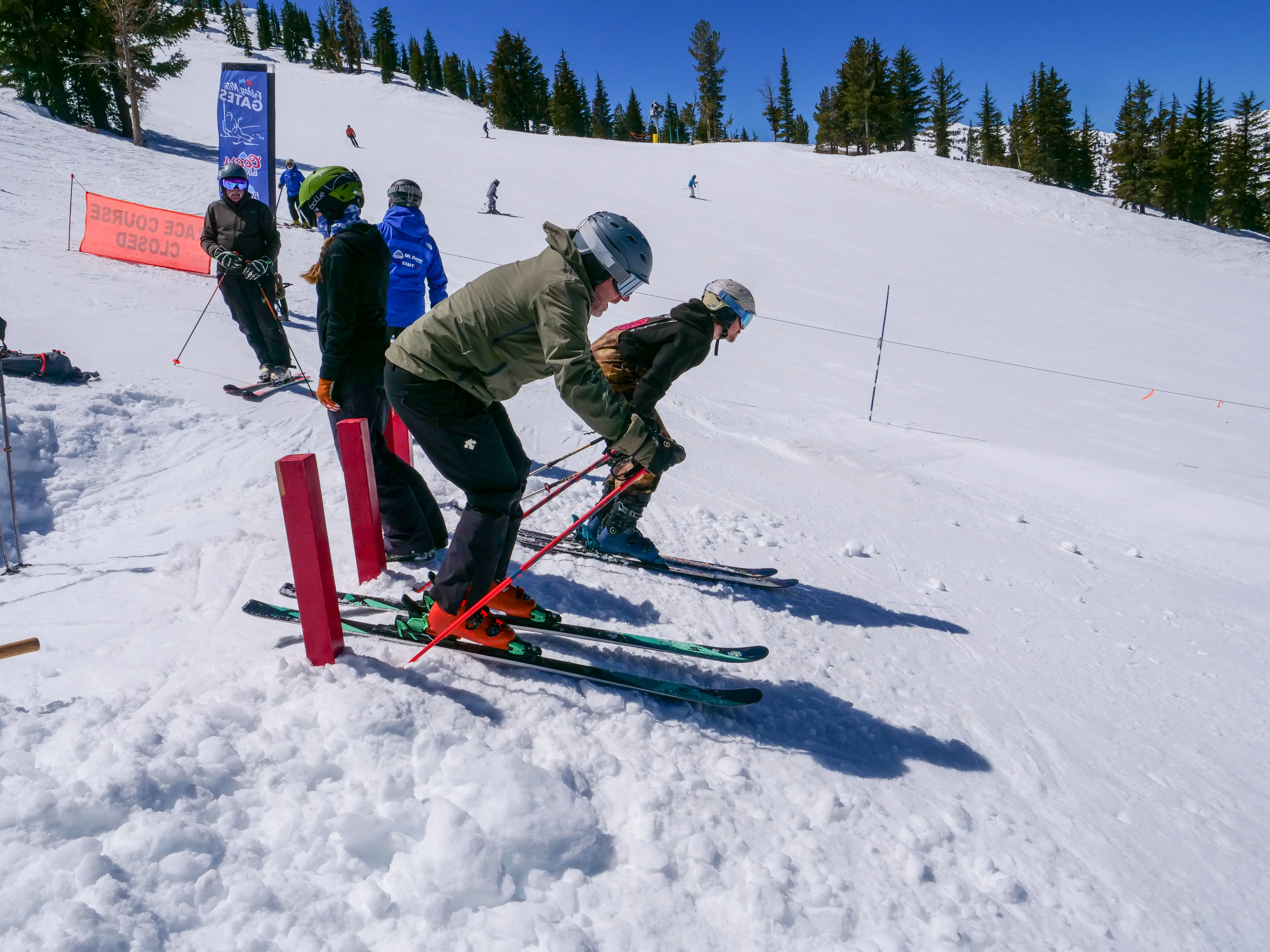 Three skiers in helmets and gear are lined up at a snowy downhill race course starting line, preparing to race. Several other people and skiers are visible in the background on the ski slope. Mt. Rose Ski Tahoe