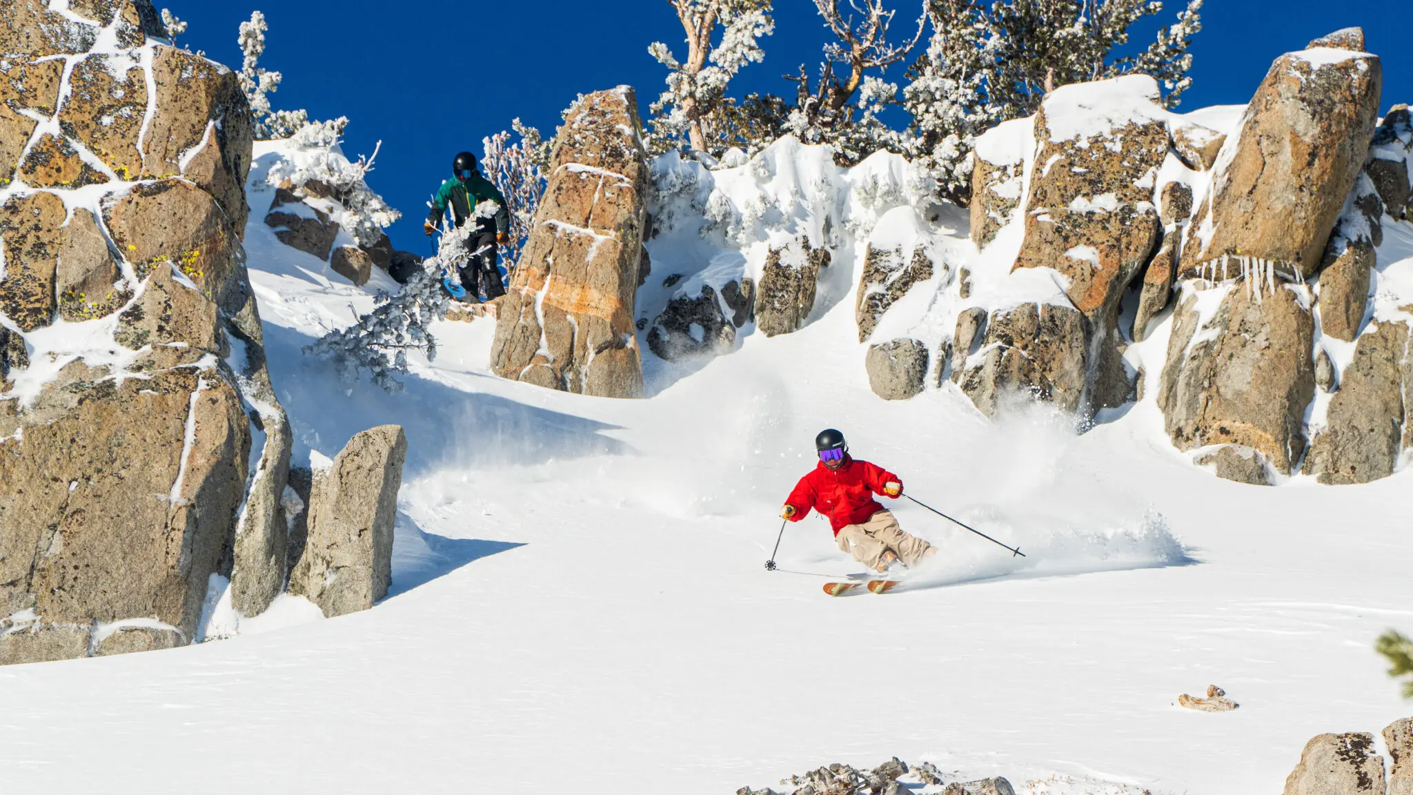 A skier in a light green jacket and black helmet moves swiftly downhill at Ski Tahoe, kicking up a spray of snow. They are surrounded by evergreen trees under a clear blue sky. Mt. Rose Ski Tahoe