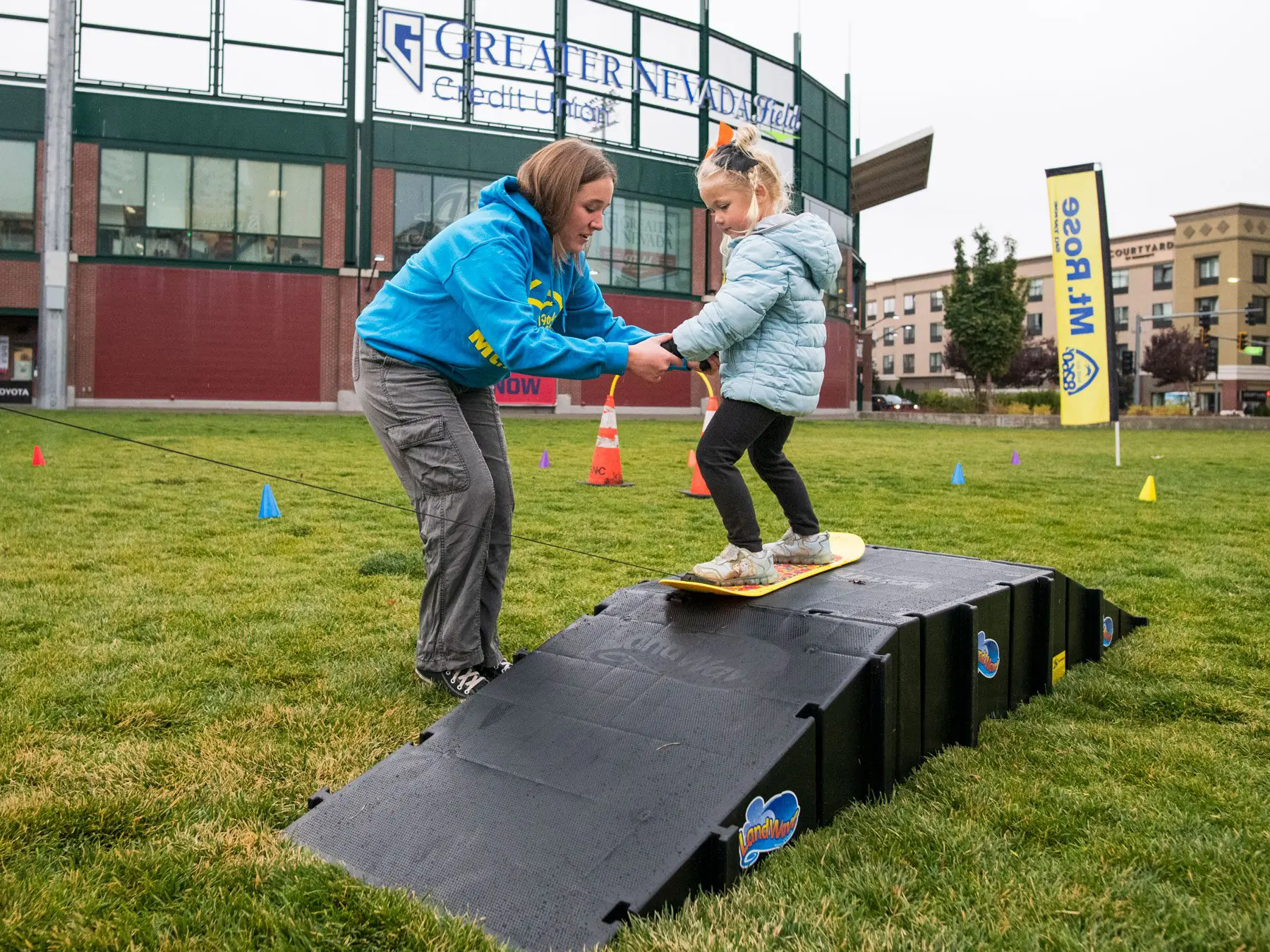 A woman helps a young girl balance on a small ramp while standing on a board, outdoors on grass near buildings and event signs. Mt. Rose Ski Tahoe