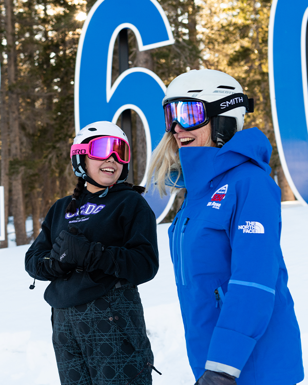 Two women wearing ski helmets, goggles, and winter clothing smile and talk while standing in the snow at Ski Tahoe, with trees and large blue numbers in the background. Mt. Rose Ski Tahoe
