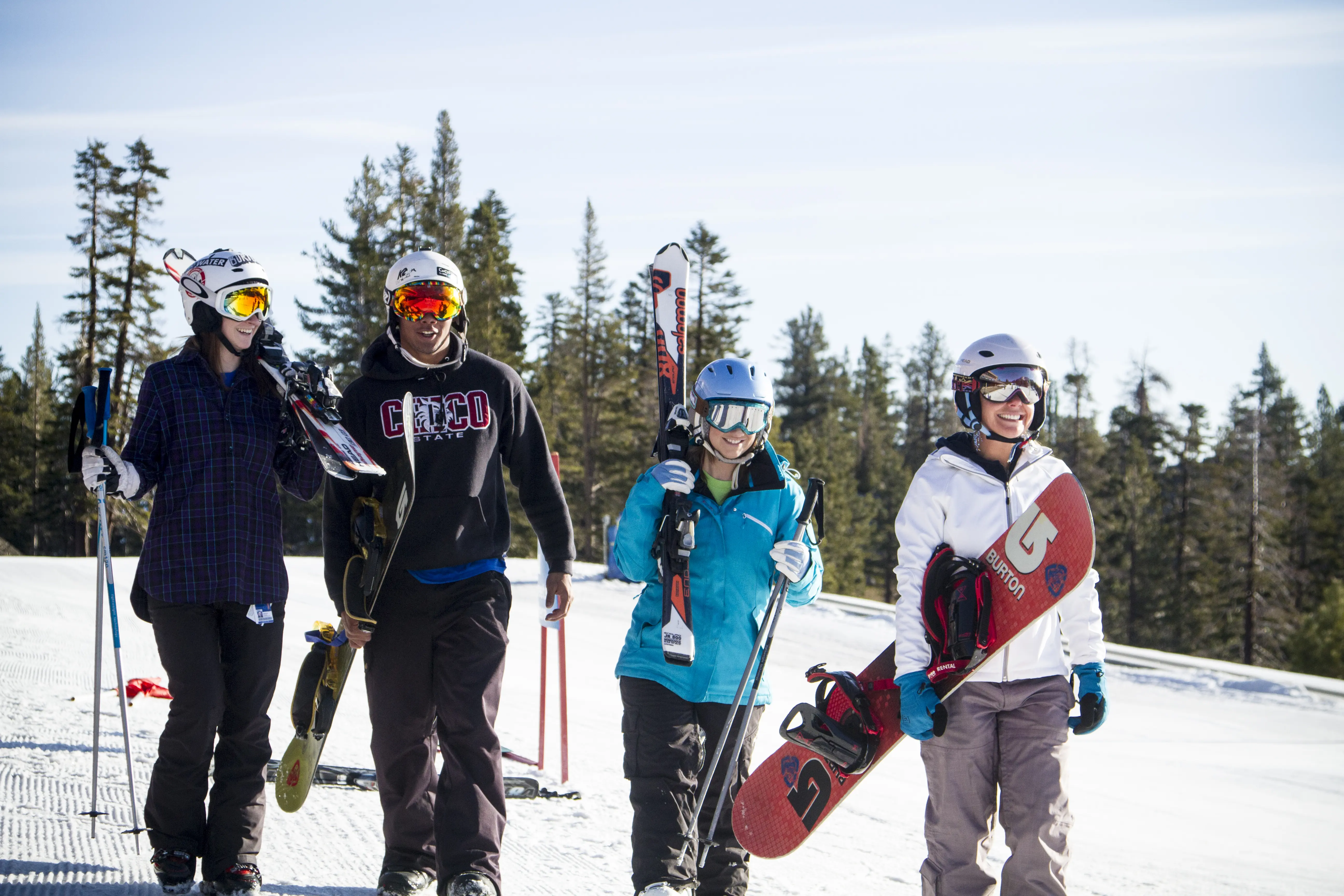 Four people in winter gear walk together on a snowy slope, carrying skis and snowboards, with trees and blue sky in the background. They appear to be smiling and enjoying the day. Mt. Rose Ski Tahoe