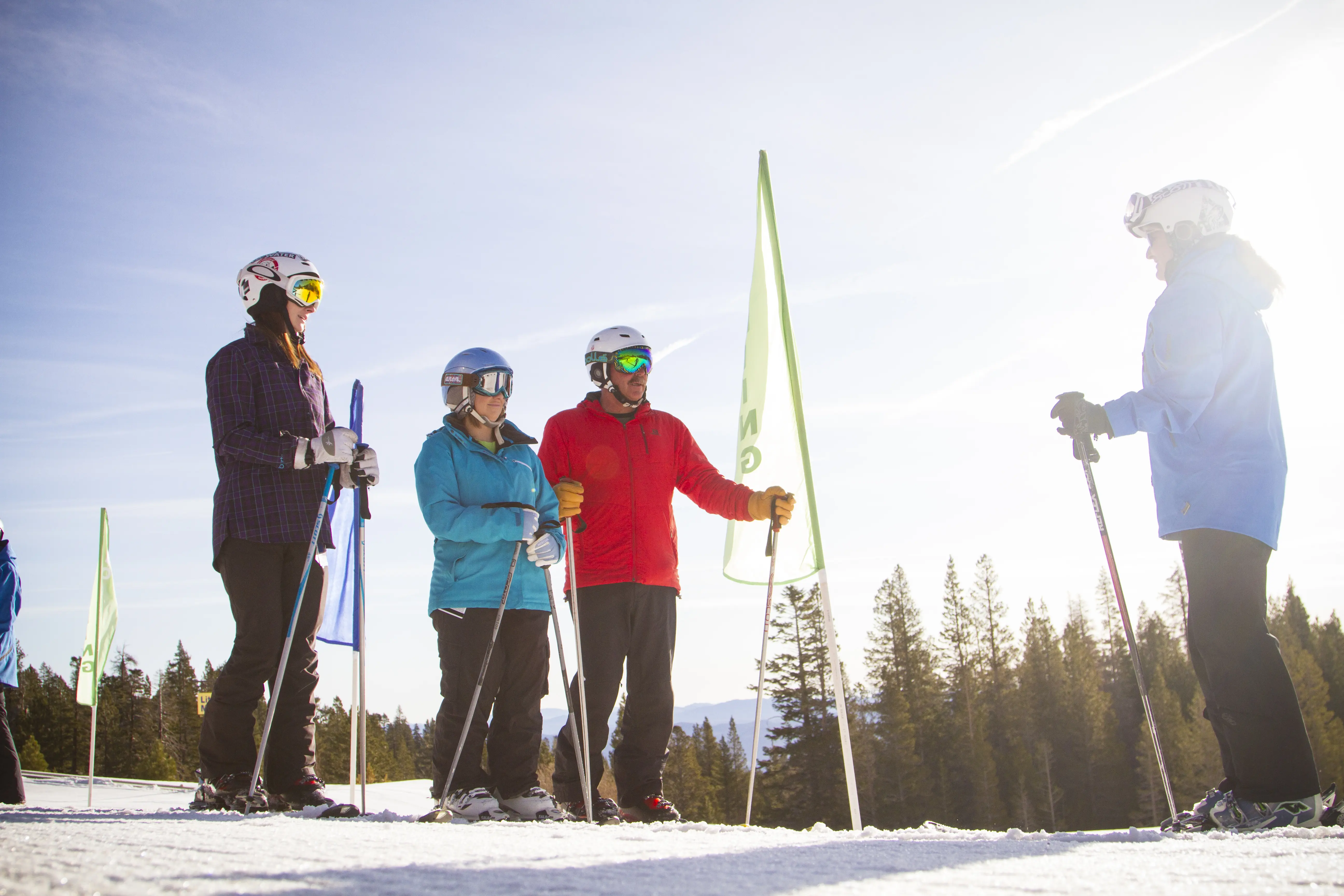 Four people in winter gear stand with skis and poles on a snowy slope, conversing in bright sunlight. Tall green flags and pine trees are visible in the background under a clear sky. Mt. Rose Ski Tahoe