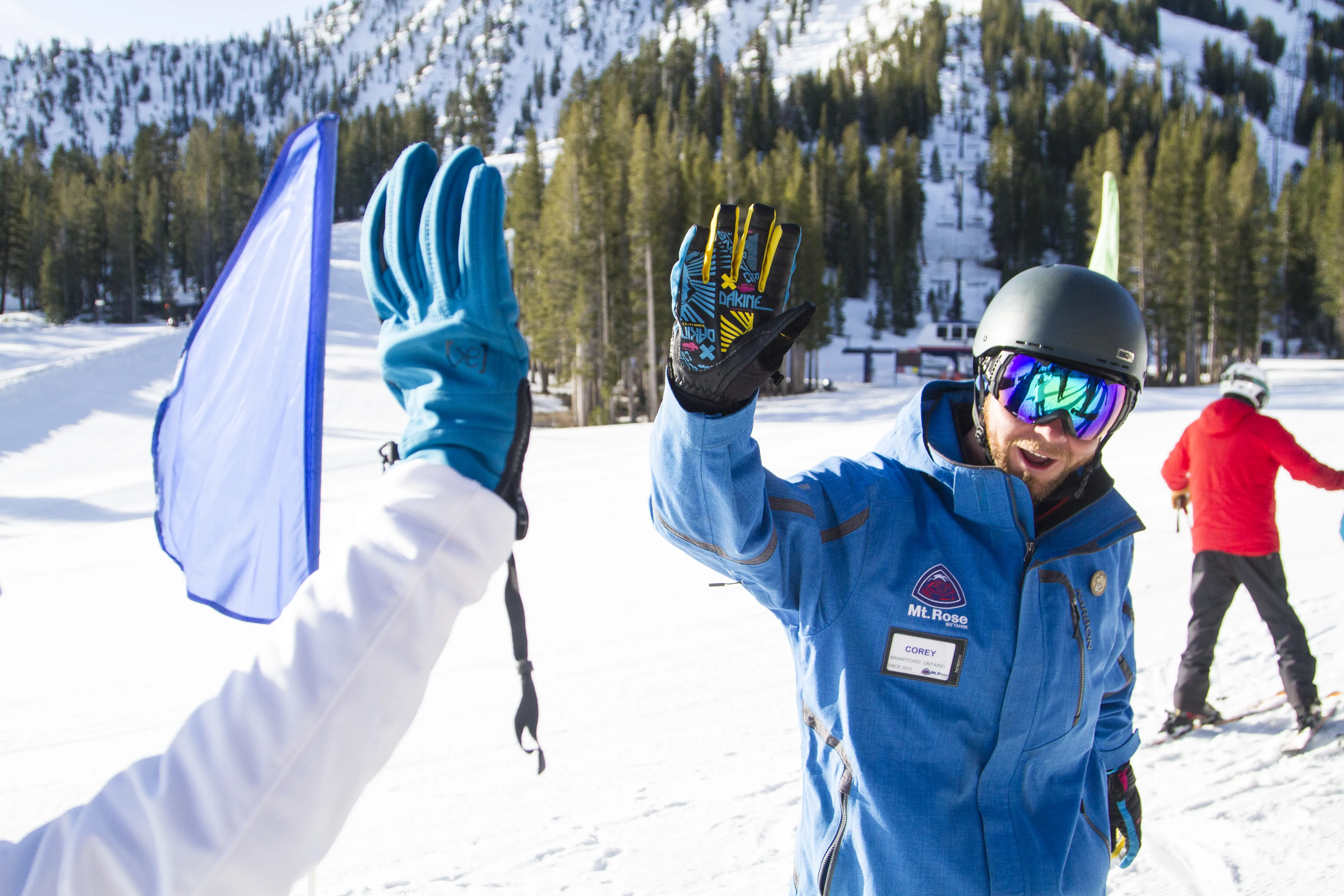 A ski instructor wearing a blue jacket and helmet gives a high-five to another person on a snowy mountain slope, with trees and skiers in the background. Mt. Rose Ski Tahoe
