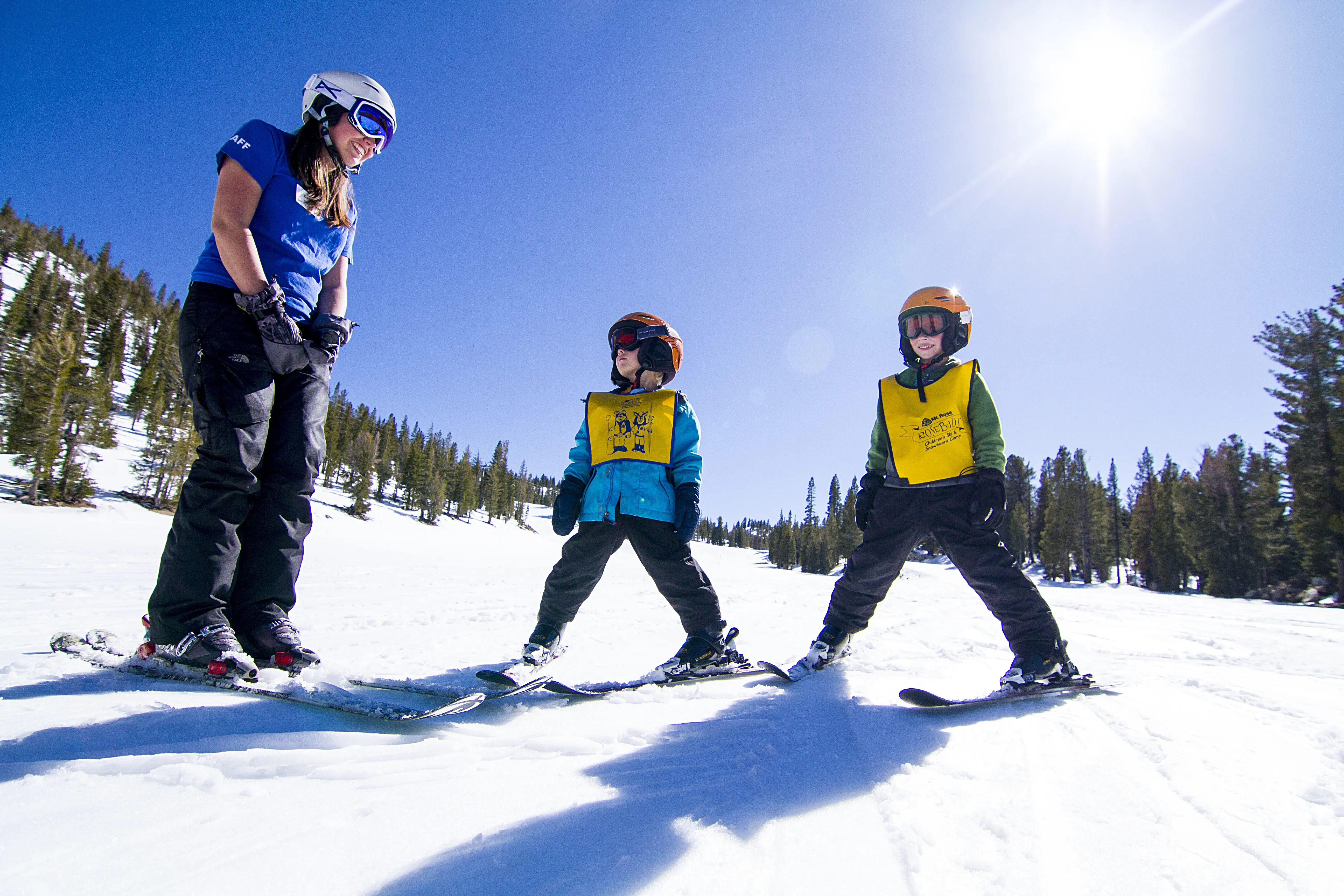 A ski instructor teaches two children in yellow vests how to ski on a snowy slope under a bright sun, with tall pine trees and a clear blue sky in the background. Mt. Rose Ski Tahoe